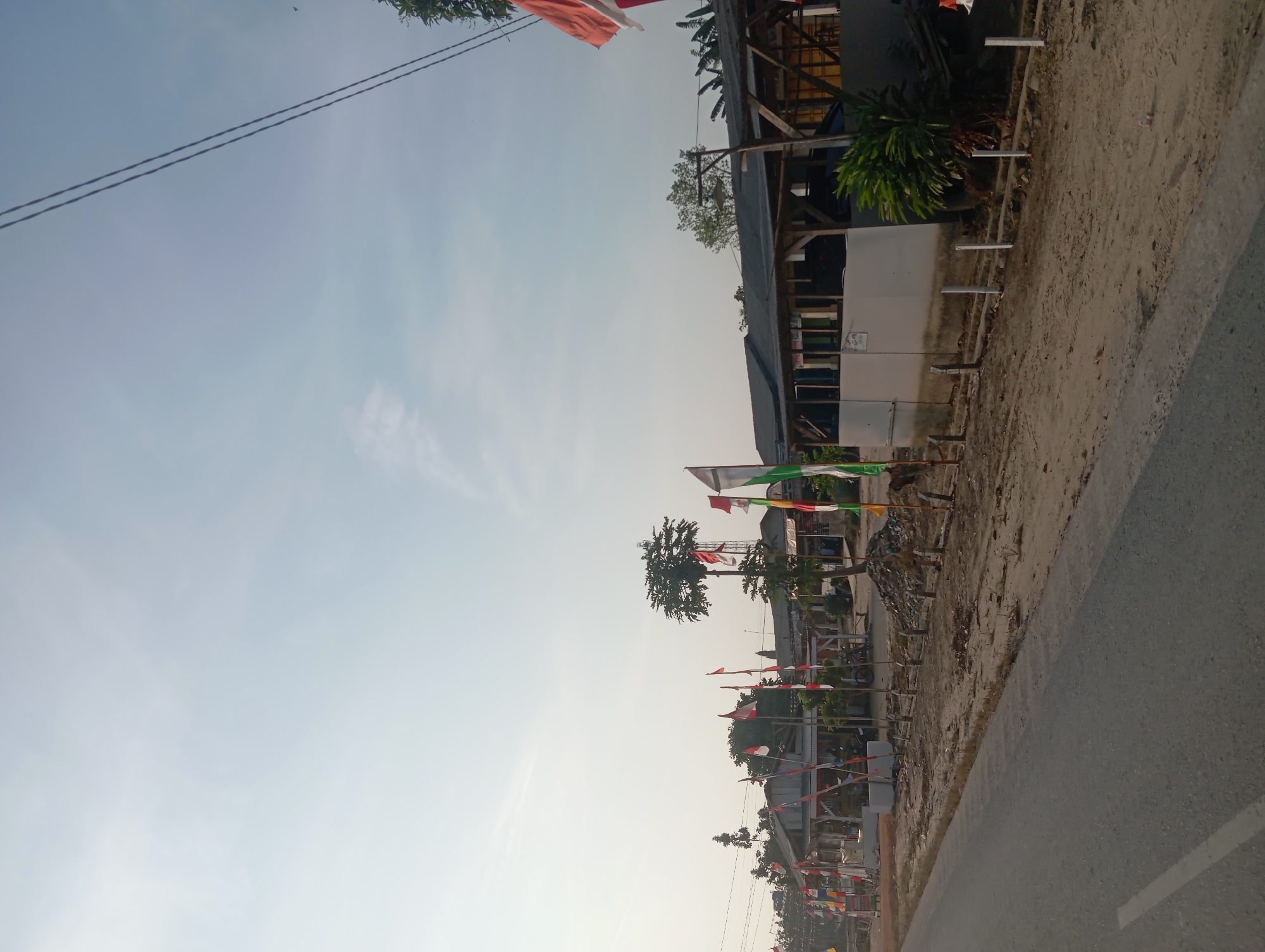 The image depicts a street scene in Dente Makmur, Indonesia, featuring a row of small businesses or stalls along the roadside. The sky is clear and pale, suggesting it might be early morning or late afternoon. Numerous Indonesian flags, predominantly red and white, are displayed on poles, indicating a national celebration or commemoration. The foreground shows a paved road with a white line marking the lane. Beyond the road, a sandy area is lined with these flags, leading up to a series of simple, open-sided structures with corrugated roofs. Some of these structures appear to be shops or food stalls. Several trees, including a distinctive one with a very full canopy resembling a pruned tree or a cell tower disguised as a tree, are visible behind the buildings. A few motorcycles are parked near the stalls. In the distance, more buildings and trees can be seen. The overall impression is that of a local commercial area decorated for a patriotic event.