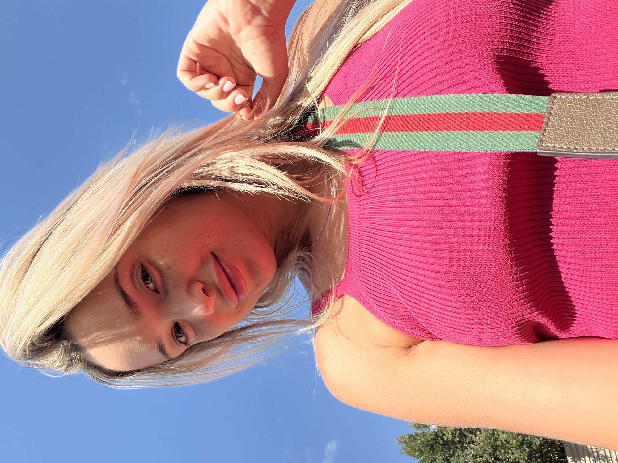 A low-angle, medium shot captures a blonde woman from the chest up, looking at the camera with a slight smile. She is wearing a vibrant pink ribbed tank top with a multi-colored strap, featuring red and green stripes, across her chest. Her hair is a light blonde, and she is holding a section of her hair with her right hand, which has neatly manicured white nails. The background is a clear blue sky with a few faint white clouds. To the left of the frame, the top of a green tree is visible, suggesting an outdoor setting. The overall lighting indicates a sunny day. The scene is set in Kryzhanivka, Ukraine.