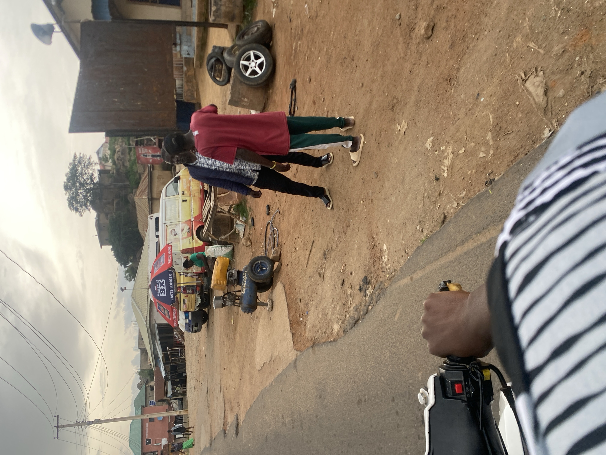 The image depicts an outdoor scene in Kabong, Nigeria, possibly a roadside market or workshop. In the foreground, a hand is visible operating a motorcycle's handlebar, suggesting the photo is taken from the perspective of a motorcyclist. The middle ground shows three individuals, two men and one woman. One man is wearing a dark blue jacket and a patterned shirt, while another is wearing a red t-shirt and green and white striped pants. The woman is wearing a green t-shirt and is seated. They appear to be interacting or observing something.
Adjacent to them is a yellow and white van with branding that includes "DANGOTE 3X LASTS LONGER YIELDS MORE." Nearby, there is a blue air compressor with a yellow tank and wheels. Several tires, including one with a silver rim, are stacked on concrete blocks to the right of the frame. The ground is unpaved, composed of reddish-brown dirt and some scattered debris. In the background, buildings with corrugated metal roofs and residential structures are visible, along with a utility pole with numerous overhead wires. The sky is overcast. The overall impression is a bustling, somewhat cluttered environment typical of a commercial area in Nigeria.