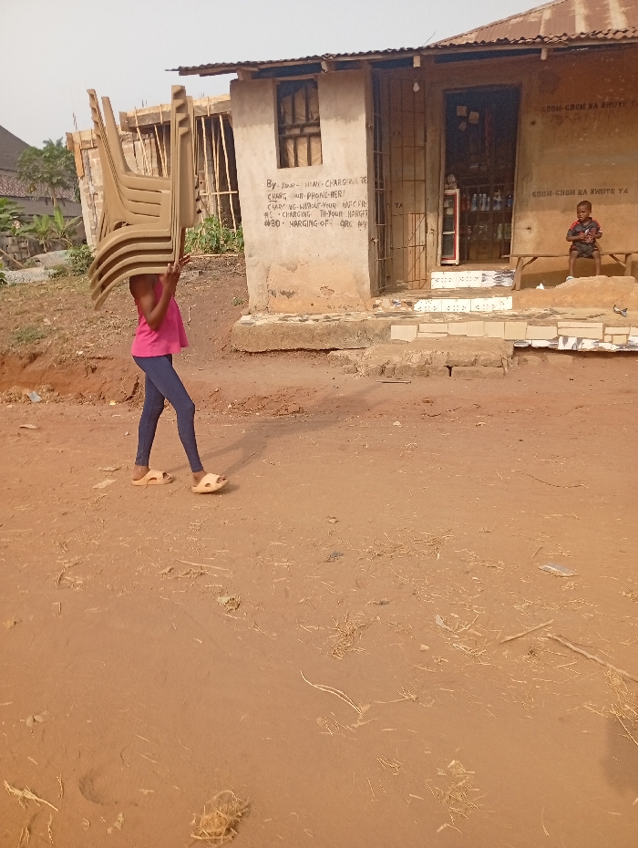 A person, likely a child, is carrying a stack of tan-colored plastic chairs on their head, walking on a dirt road. In the background is a weathered building with signage indicating phone charging services and prices. A young boy sits on a bench in front of the building. The ground is dirt-covered with sparse vegetation.