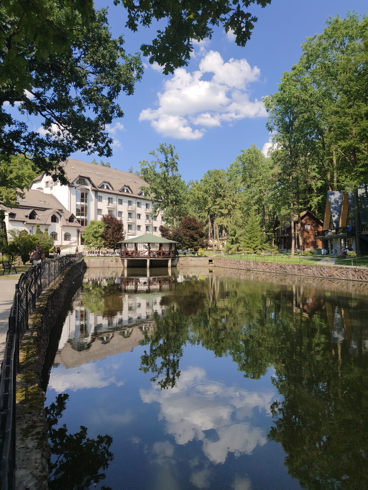 The image depicts a serene waterside view of a resort-like complex, likely located in Solotvyno, Ukraine, known for its mineral lakes and recreational facilities. The scene is dominated by a calm, dark body of water reflecting the surrounding environment. On the left, a multi-story building with a light-colored facade and a brown tiled roof stands prominently. It features multiple windows and balconies, suggesting it's a hotel or accommodation. In front of this building, a covered wooden pavilion with a green roof is situated over the water, possibly a dining or relaxation area.
To the right of the main building and across the water, several smaller wooden cabins or houses are visible, nestled amongst lush green trees. These structures have a rustic architectural style, with dark wood and angular roofs. The water's edge is lined with a decorative stone wall and a black metal fence along the paved walkway on the left.
The environment is characterized by abundant greenery, with mature trees framing the buildings and lining the background. The sky above is a bright blue, dotted with fluffy white clouds, which are also clearly mirrored in the still water. A few people can be seen walking along the path on the left, and one person is seated on a bench. The overall impression is one of tranquility and natural beauty, a place designed for leisure and relaxation.