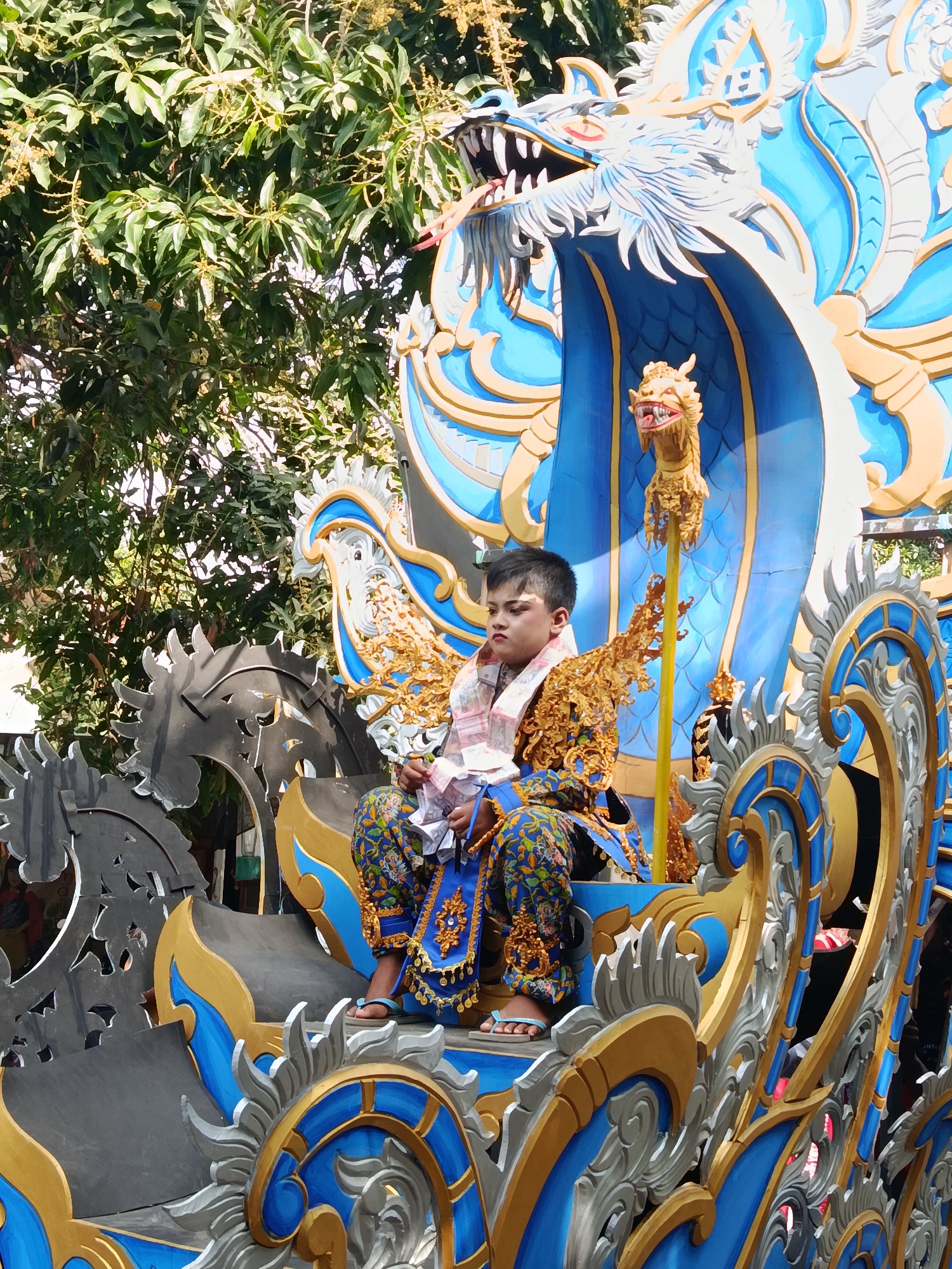 In the city of Ketowan, Indonesia, a young boy is seated on a decorative float during what appears to be a parade or cultural event. The float is ornately designed, featuring a large blue and gold dragon motif, with the boy positioned in front of its head. He is wearing traditional, elaborate clothing in shades of blue, gold, and white, adorned with intricate patterns and embellishments. The boy is holding what appears to be money in his hands and looking to the side. In the background, lush green trees suggest an outdoor setting, possibly a street or public square. The overall scene is festive and vibrant, indicative of a celebration.