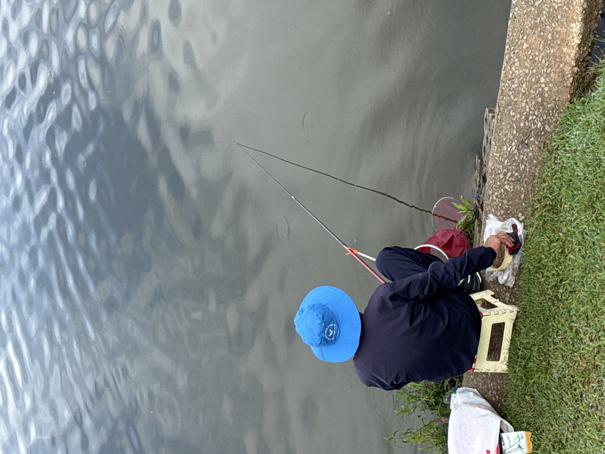 An adult sits on a small, folding, yellow stool at the edge of a body of water, likely a lake, with two fishing poles cast into the water. The individual is wearing a blue bucket hat and a navy long-sleeved shirt with dark pants. A maroon bag sits next to the person near the edge of the lake. To the person's right, on the grassy shoreline, is a white plastic bag and some food packaging. The shoreline is concrete with grass and foliage adjacent to it. The water has a rippled surface. The location is Da Lat, Vietnam.