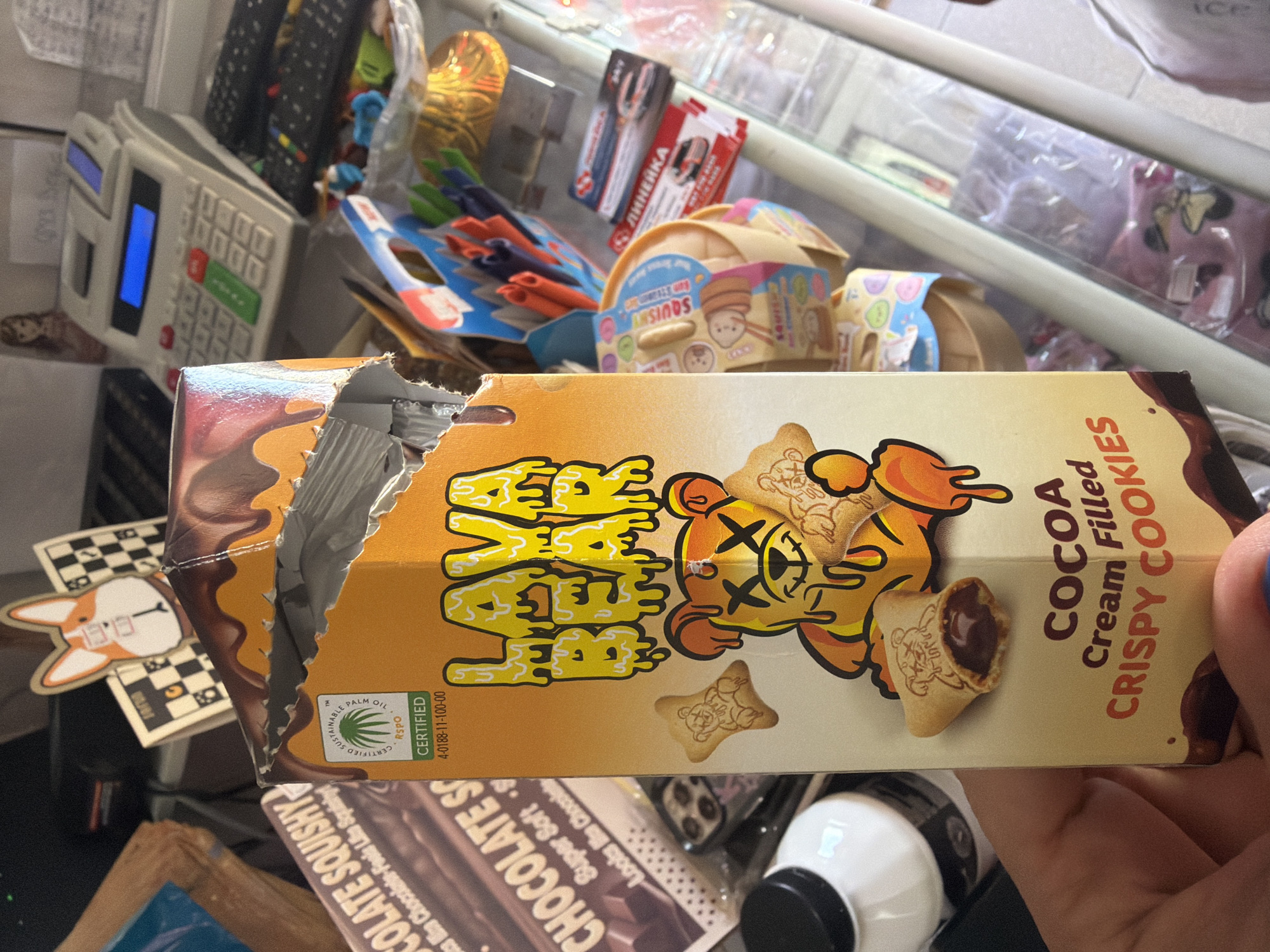 The image shows a close-up of a hand holding a damaged box of "Lava Bear" cocoa cream-filled crispy cookies. The box is predominantly orange and yellow with a melting chocolate design and a cartoon bear graphic. The cookies are shaped like bears and appear to have a chocolate filling. In the background, there are various objects on a shelf, including a calculator, remote controls, and other food packaging. The scene appears to be indoors, possibly in a store or home environment. There is no identifiable information in the image that would link it to the city of Ravno pole, Bulgaria.