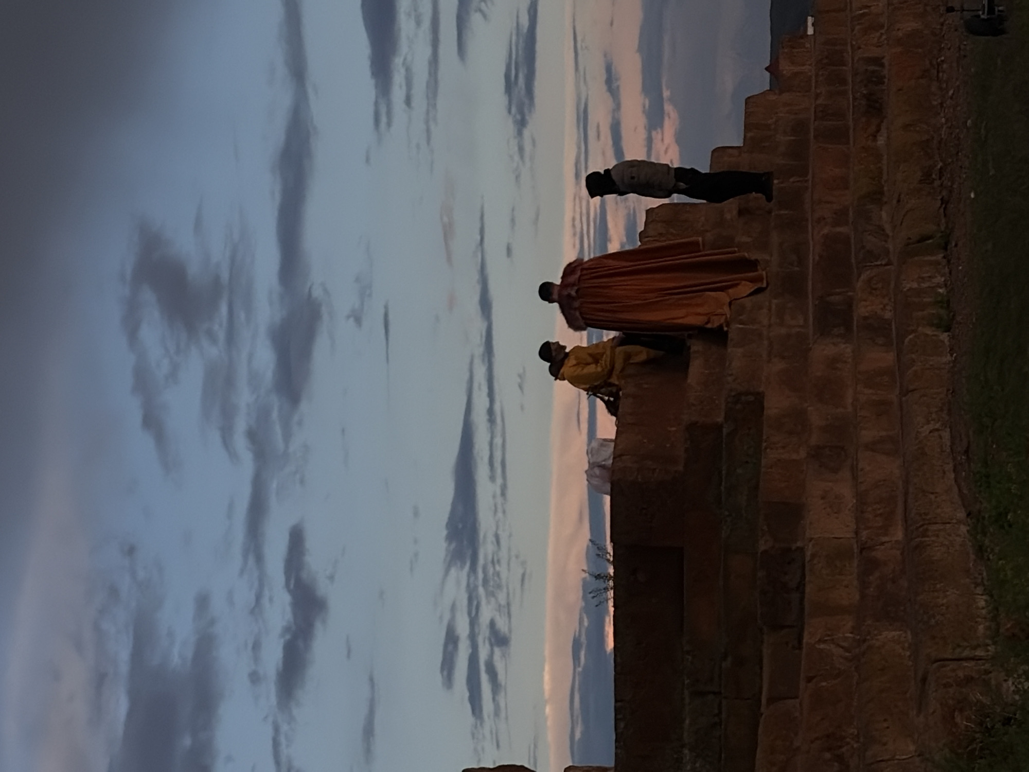 The image shows three people standing on a large stone structure resembling stairs or terraces. The sky in the background is filled with clouds, with a blend of blues, grays, and a hint of pink near the horizon. The stone structure appears to be old, with a weathered and uneven surface.
From the bottom of the structure to the top, one person is sitting down while the other is standing up wearing a long orange robe. Lastly, there's another person wearing darker clothing standing alone on a higher level. Vegetation is visible on the ground to the right of the structure. There is an object behind the person on the top step, possibly a camera on a tripod.
Given the reference to Anipemza, Armenia, this could be a part of the archaeological site of Ani, a ruined medieval Armenian city known for its stone architecture. However, without further context, it is impossible to confirm this definitively.