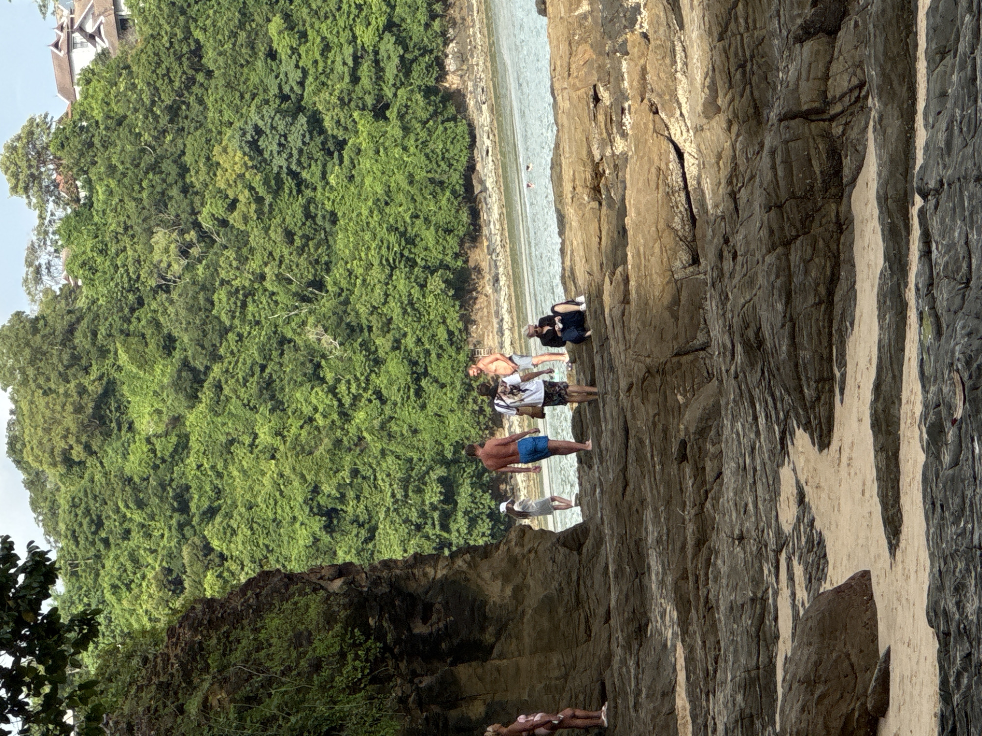 The image captures a rocky shoreline with a lush, tree-covered hill in the background. Several people are present on the rocks and near the water's edge. On the left, a woman in a pink swimsuit stands facing away from the camera. Further to the right, a group of three individuals walks along the shore. One man, wearing blue swim trunks, is facing away. Another person, in a white top and dark bottoms, walks ahead of him, while a third man in a floral print shirt and shorts is seen from behind. In the foreground, a person wearing a black top and dark pants is seated on the rocks, looking down. The sea is calm and appears to be a pale blue-green. The rocky terrain is varied, with some areas of exposed sand. Buildings are visible atop the hill in the distance, nestled amongst the dense greenery. The scene suggests a tropical coastal environment, likely in the city of Wichit, Thailand, given the typical landscape of the region.
