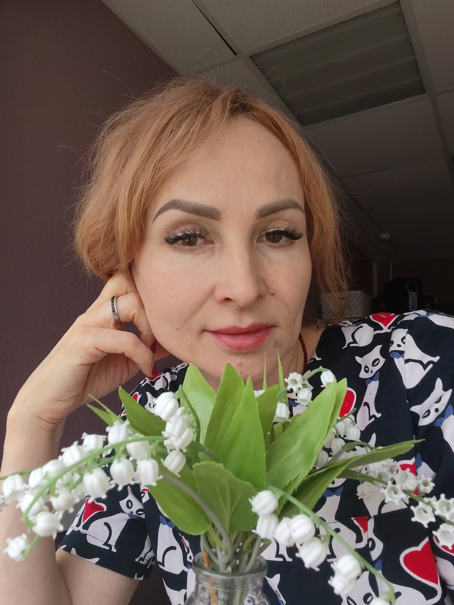 The image is a close-up shot of a woman with reddish-blonde hair posing with a small bouquet of lilies of the valley. The woman has fair skin, defined eyebrows, and is wearing makeup, including false eyelashes and lipstick. She is wearing a ring on her left hand and a patterned black, white, and red top. Her left hand is resting on her face near her ear. The lilies of the valley are in a clear vase in the foreground. The background includes a maroon-colored wall on the left and a drop ceiling with fluorescent lighting towards the top right. The location is the city of Chaikovsky, Russia.