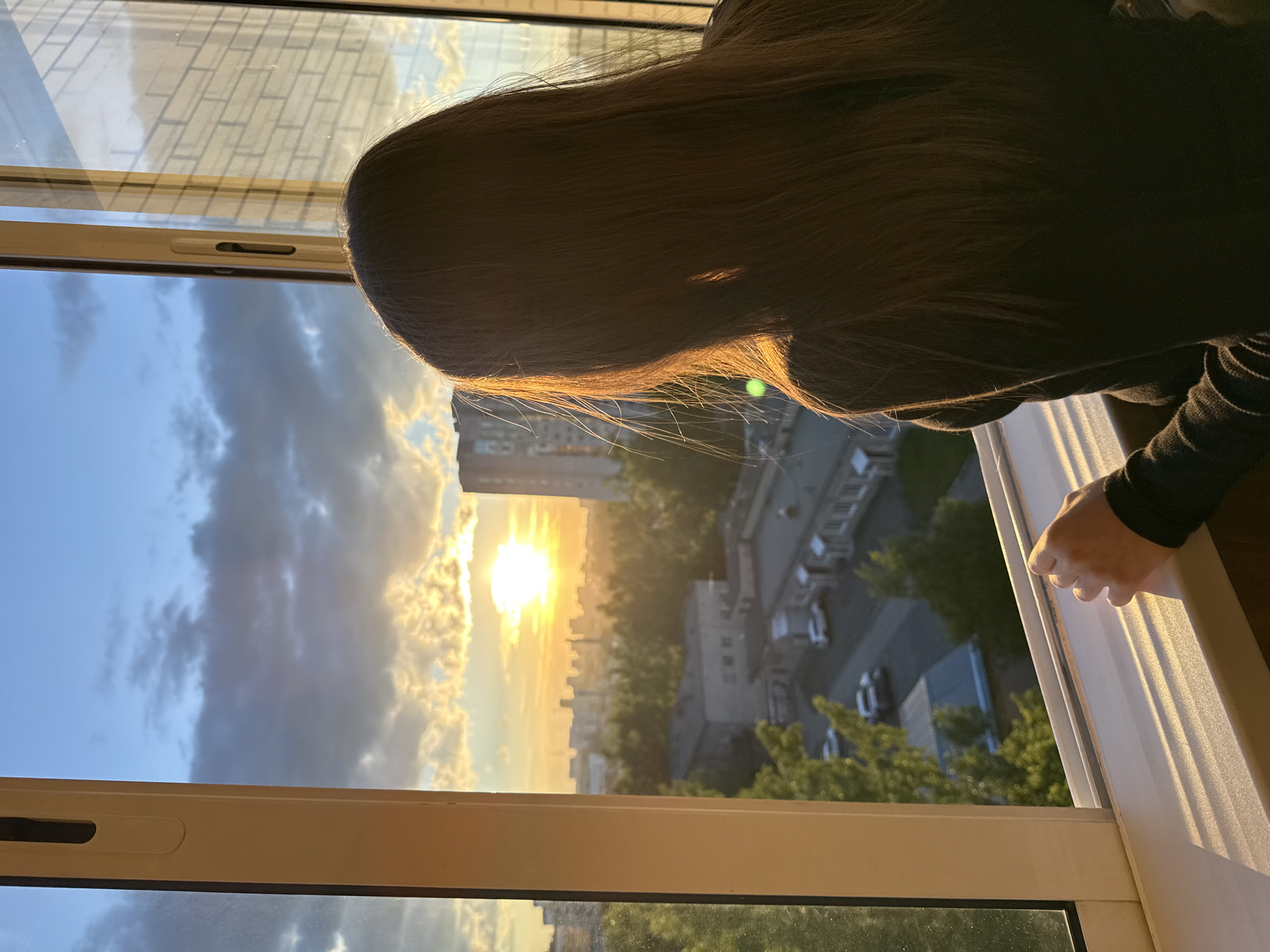 An overhead view captures a person with long, dark hair, seen from behind, leaning out or looking through a window. The golden light of the setting sun backlights their hair, creating a bright halo effect. Their right hand is visible, resting on the white window frame.
Through the window, a vivid sunset unfolds over an urban landscape, likely in Izhevsk, Russia. The sun, a brilliant orb of intense orange and yellow light, is positioned low on the horizon, casting a warm glow across the scene. The sky is partly cloudy, with dramatic darker clouds contrasting with lighter areas illuminated by the sun's rays.
Below, a sprawling city or suburban area is visible, featuring numerous buildings of varying heights, interspersed with green trees. Roads and parked cars can be discerned in the distance, indicating an active settlement. The perspective suggests a vantage point from a higher floor of a building. The image conveys a serene moment of observation during golden hour. No visible text is present.