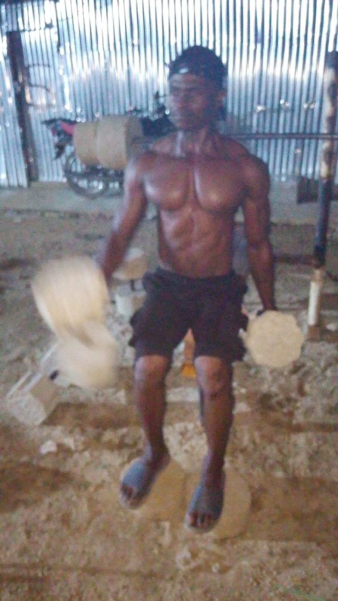 A shirtless Black man wearing dark shorts and blue flip-flops is seated on an overturned bucket, holding a round, off-white object in each hand. His body is positioned facing forward and slightly to the right. The man's musculature is visible. His hands are blurred due to motion. The background features a corrugated metal wall and the blurred image of a motorcycle with a tan cargo box. The ground appears to be dirt and debris. The scene is dimly lit, suggesting an indoor or covered outdoor space. The location is identified as Indo, Nigeria.