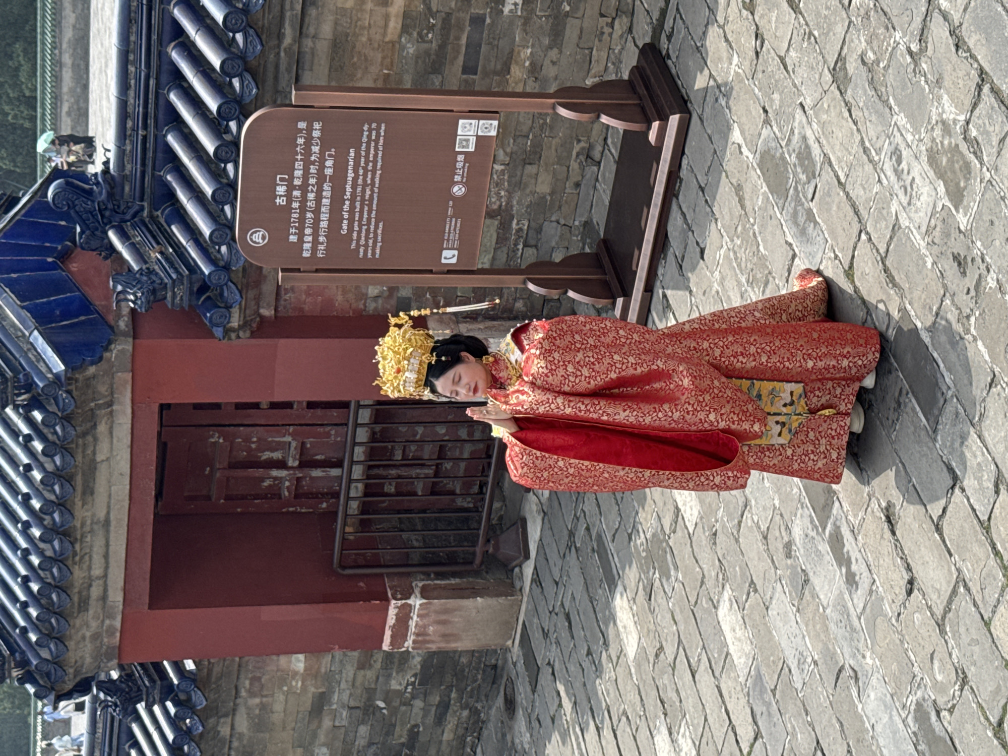 The image shows a woman in traditional Chinese dress, kneeling in what appears to be a courtyard in Dongcheng District, China. She wears an elaborate golden headdress and a red patterned robe. Her hands are clasped together in front of her. Behind her is a red building structure with a dark blue tiled roof. To the side of the woman is a sign in both Chinese and English, marking the location as the "Gate of the Septuagenarian," which was built in 1781 to reduce the amount of walking required for Emperor Qianlong. The courtyard is paved with grey stone bricks.
