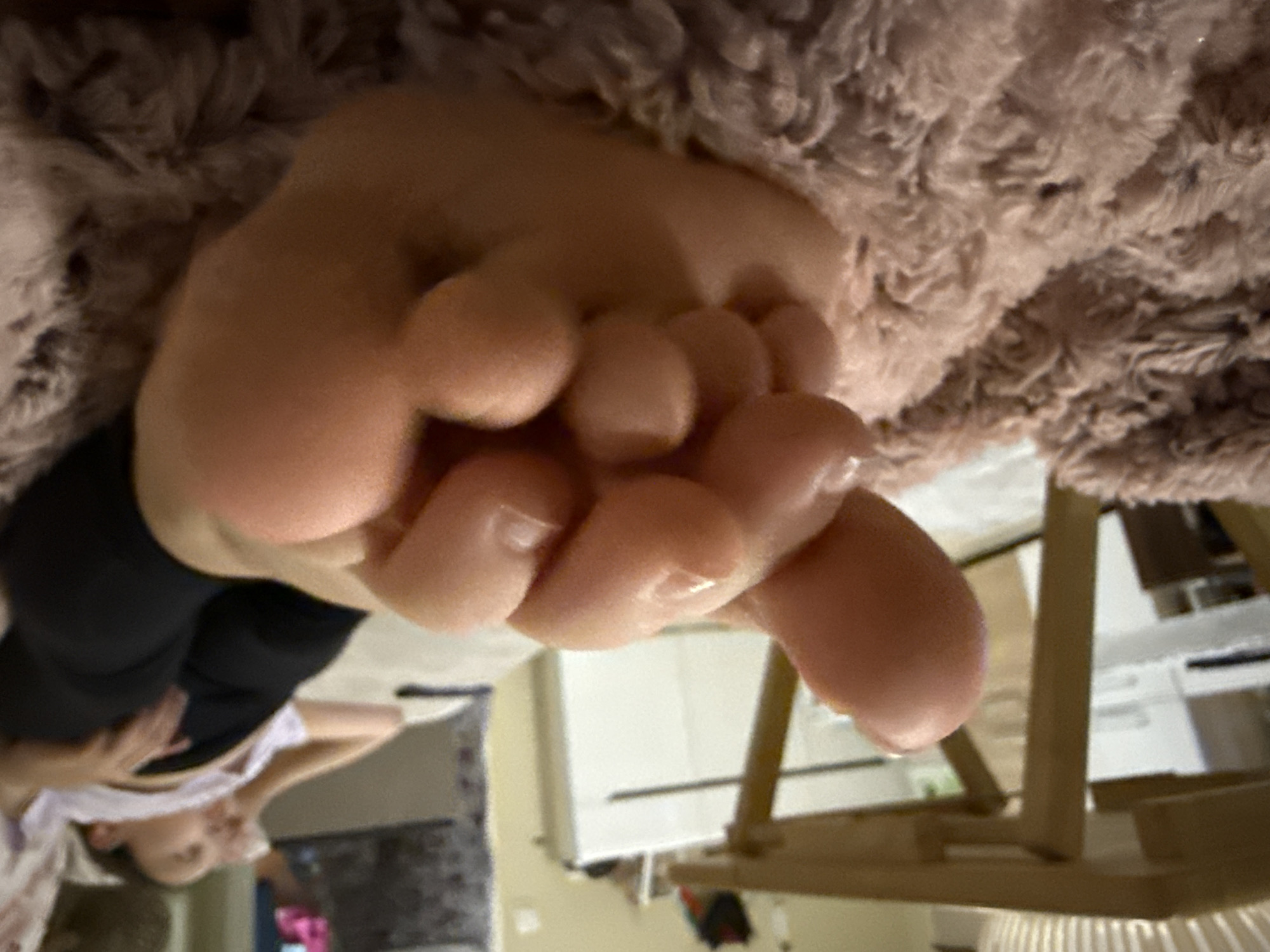 The image presents an interior scene with a close-up, low-angle perspective focused on a bare human foot resting on a textured, light brown/pinkish plush surface, likely a blanket or rug. The foot is oriented with the sole partially visible and the toes slightly curled towards the viewer, showing visible toenails on the smaller digits. The skin appears moisturized and free of blemishes.
In the mid-ground, a second individual is visible lying down and out of focus on a light-colored surface, possibly a bed or sofa. This person is wearing black leggings and a white or light-colored top. Their head is resting on a pillow or armrest, and one hand is positioned near their torso. Their posture suggests relaxation or rest.
The background is significantly blurred, revealing elements of an indoor environment. To the left, a light-colored wooden table with slatted detailing is partially visible. Further back, a white piece of furniture, possibly a cabinet or dresser, stands against a wall. A dark, indistinct object rests on top of this furniture. The overall lighting suggests an artificial indoor light source or a dim ambient light.
The specific location in the city of Targovishte, Bulgaria, cannot be identified from the visual information presented in the image.