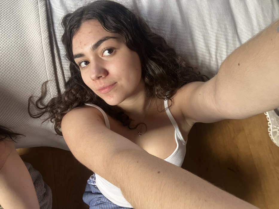 A young woman with curly brown hair takes a selfie, lying down on what appears to be a striped cushion. She wears a white tank top and blue striped bottoms. Her arm is extended toward the camera, showcasing visible arm hair. A necklace is visible around her neck. Part of another person and white sneakers are seen in the peripheral.