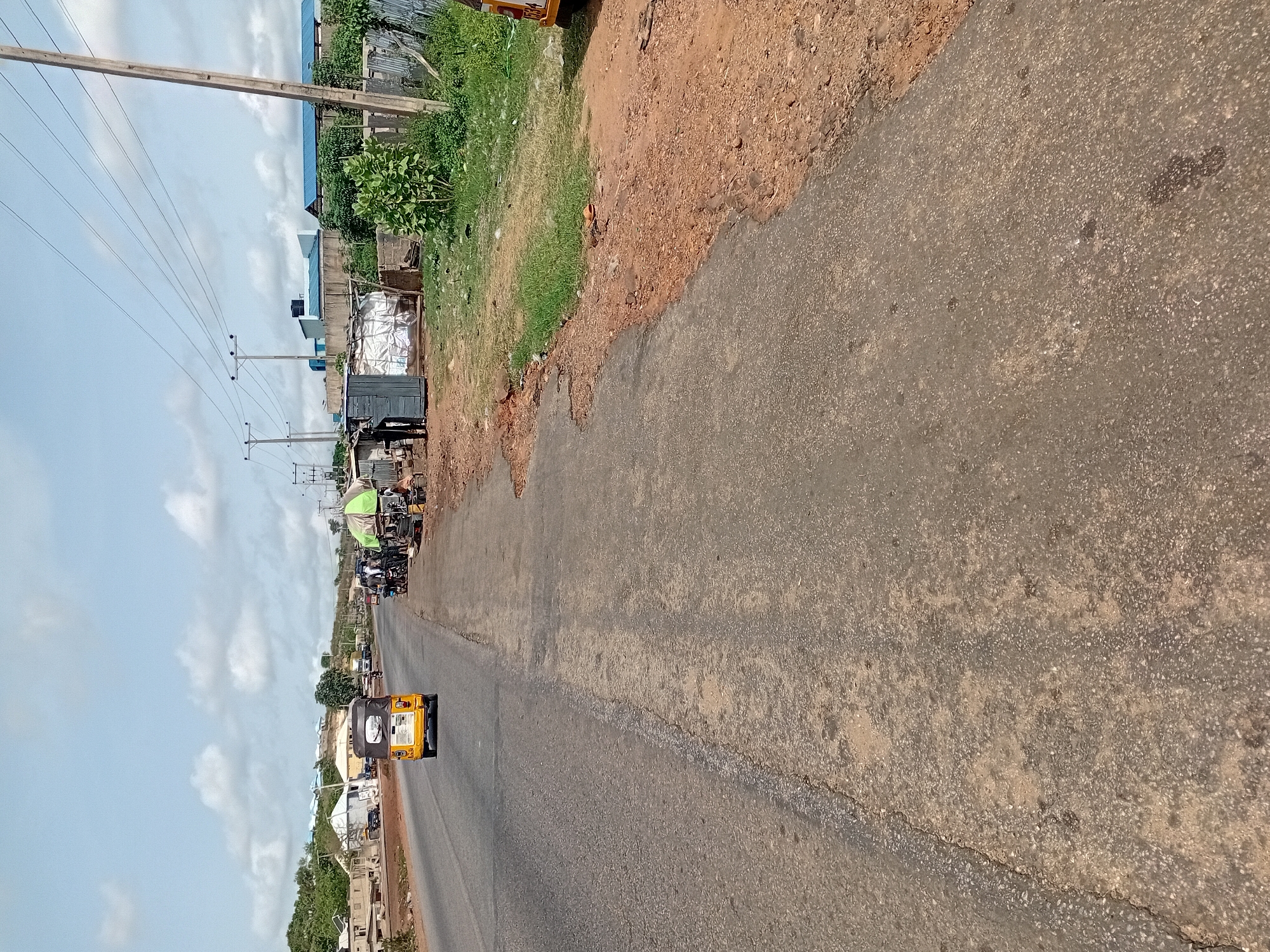 The image displays a daytime street scene along a road in Lokoja, Nigeria, captured under a bright, cloudy sky. The asphalt road, showing signs of wear with cracks and uneven patches, extends from the foreground into the distance, appearing to be a two-lane thoroughfare without clear markings.
To the left of the road, a narrow dirt and grass shoulder borders the pavement, behind which stand various buildings that could be commercial structures or informal market stalls, interspersed with scattered vegetation. Numerous utility poles with power lines run alongside the road on this side. On the right, a wider, unpaved shoulder composed of reddish-brown earth and sparse grass lines the road, with more distant buildings visible beyond it.
A yellow "Keke Napep" (auto-rickshaw) is a central subject in the mid-ground, heading away from the viewer. On its rear, a sticker or sign contains partial text, including "Z" followed by indistinct letters, then "NAPE", and potentially "24HRS" along with what appears to be a phone number or registration details. A partial license plate or identification number, "0584", is also visible on the upper right side of the Keke. Further down the road, several other vehicles, including what appears to be a truck and smaller vehicles or motorcycles, are visible, either moving or parked. A few indistinct figures of people can be vaguely discerned near the buildings on the left, suggesting routine daily activity.
The scene is brightly lit, indicative of morning or early afternoon, with an overcast sky preventing strong shadows. The overall setting reflects a common urban or semi-urban environment in Nigeria.
