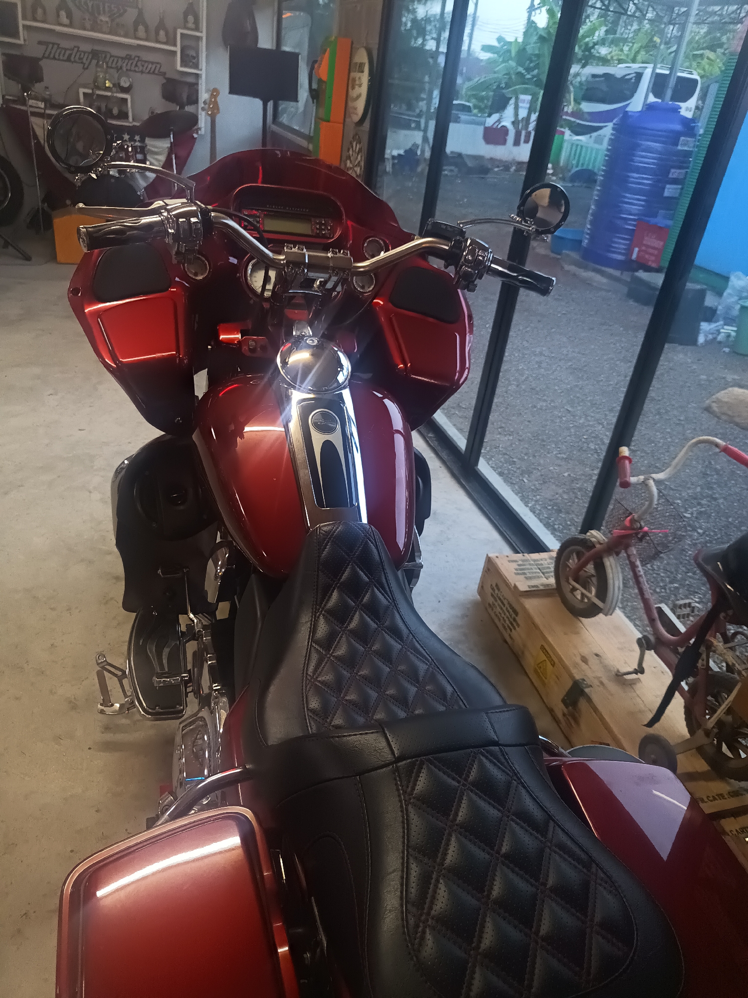 The image shows a red Harley-Davidson motorcycle parked indoors, likely in a garage or showroom. The motorcycle has a large fairing, a quilted leather seat, and chrome accents. In the background, there is a wall with Harley-Davidson branding and various decorative items. To the right of the motorcycle, there is a window showing an outdoor scene. There is also a small red bicycle next to the motorcycle. Based on the surrounding details such as the interior design and the presence of foreign language signs, it's plausible to hypothesize this image was captured in the city of Surat Thani City Municipality, Thailand.