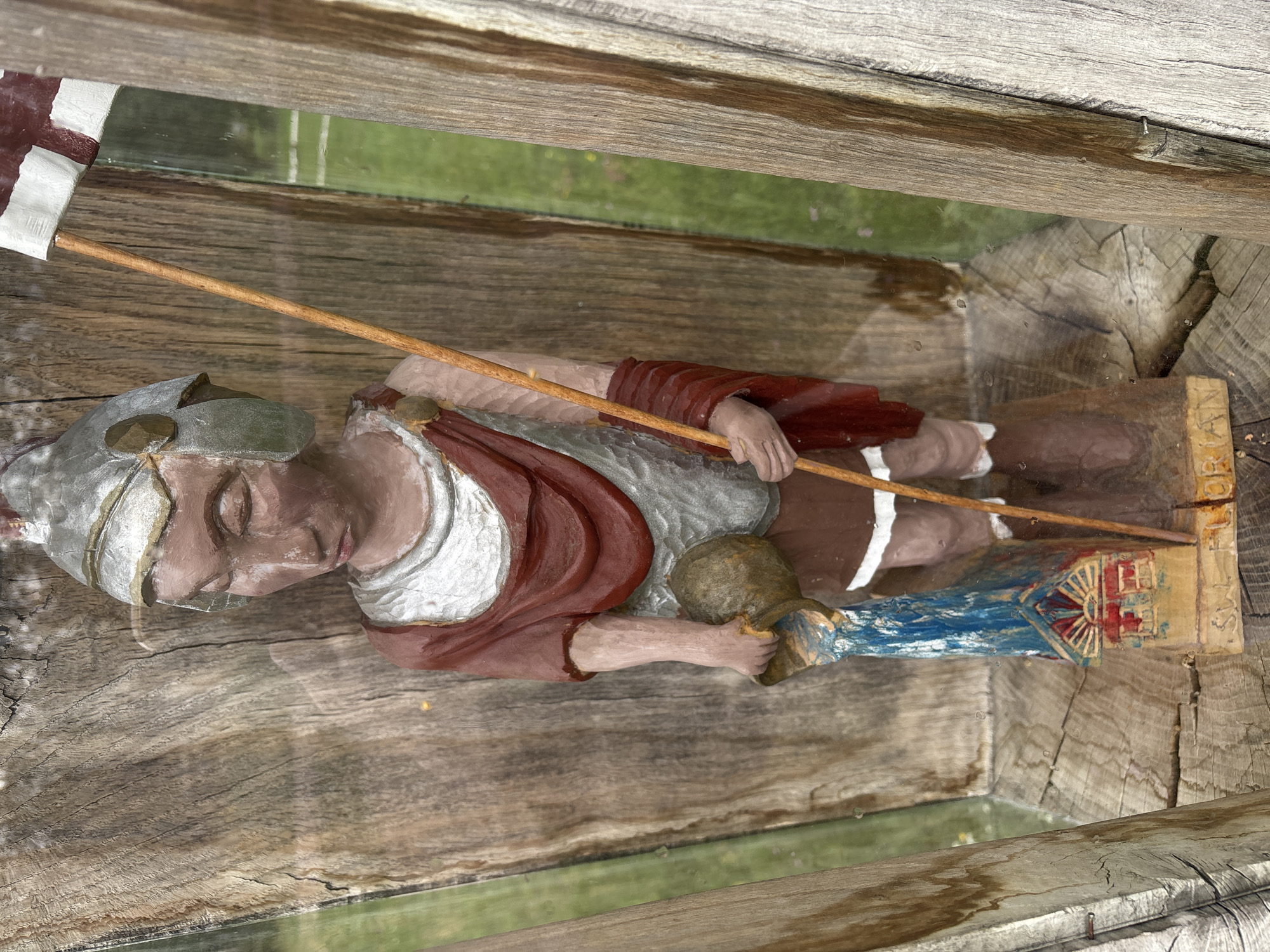 A carved wooden statue of a figure resembling a Roman soldier stands in a display niche. The figure is painted with a brown face, silver armor, a red draped garment, and a brown skirt. It holds a wooden staff with a white and red flag at the top in its right hand and a jug from which blue liquid is being poured in its left hand. At the base of the statue is a small, colorful depiction of a building with red and blue details. The statue is positioned against a weathered wooden background. The display niche is framed by rough-hewn wooden posts, and a reflective surface, possibly glass, is visible in front of the statue, showing the surrounding environment as a blurred green landscape with white lines, suggesting a field. The overall impression is that of an outdoor display, likely in a public space within Sielachowskie, Poland.