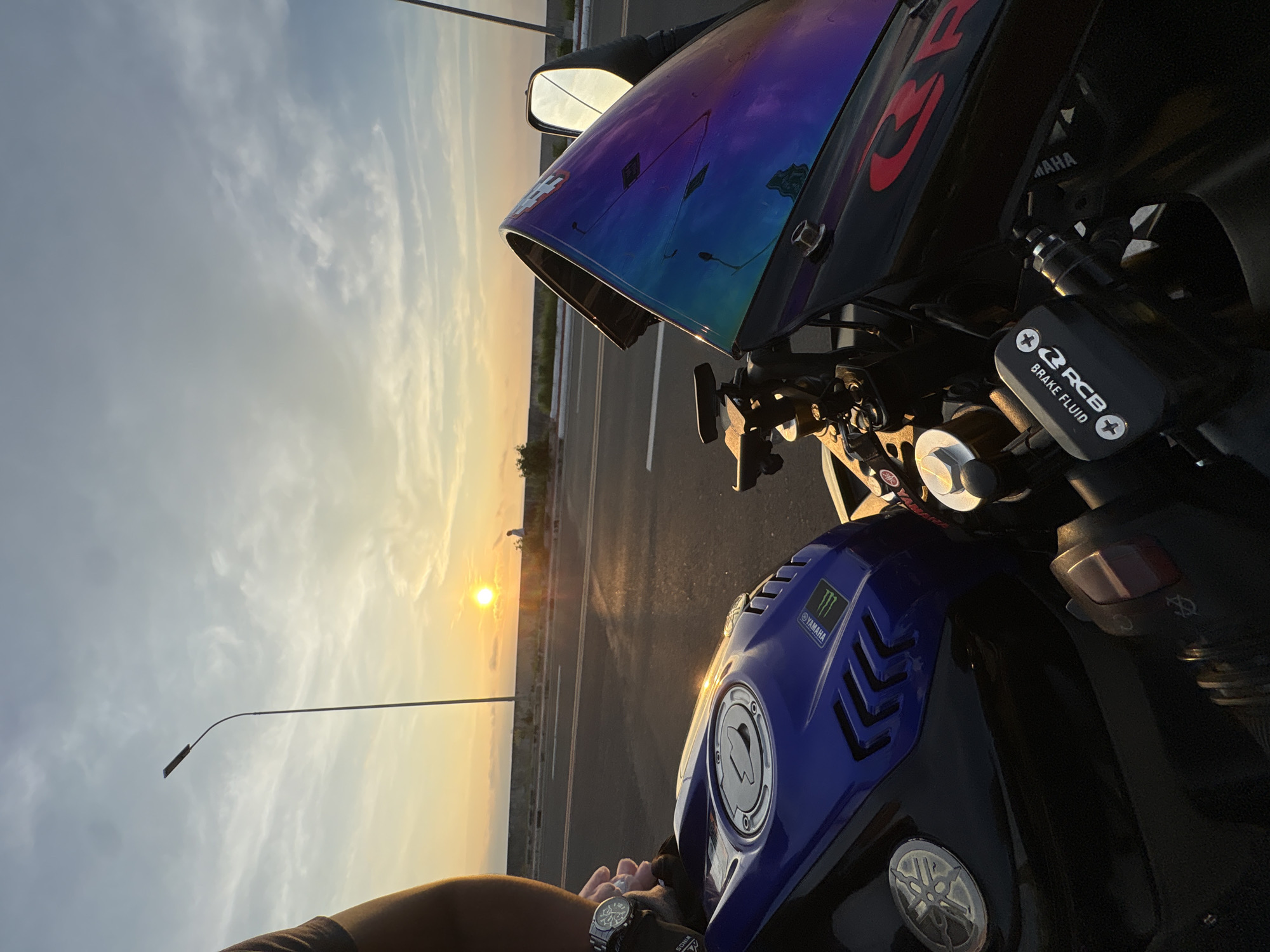 The image shows a close-up perspective of a motorcycle parked on the side of a road during sunset. The motorcycle is black and blue, with a prominent Yamaha logo on the fuel tank. The windshield of the motorcycle is iridescent, reflecting shades of purple and blue. In the background, the sun is setting on the horizon, casting a warm glow across the sky and the road. A single street lamp stands tall on the left side of the image. A person, wearing a watch on their left wrist, is partially visible on the left side of the frame, holding onto the motorcycle. The road appears to be a coastal highway, with a low concrete barrier and some vegetation visible in the distance. A person is seen walking in the far background. The scene suggests a location along the coastal road in Davao City, Philippines, likely near the Roxas Avenue stretch, known for its sunset views.