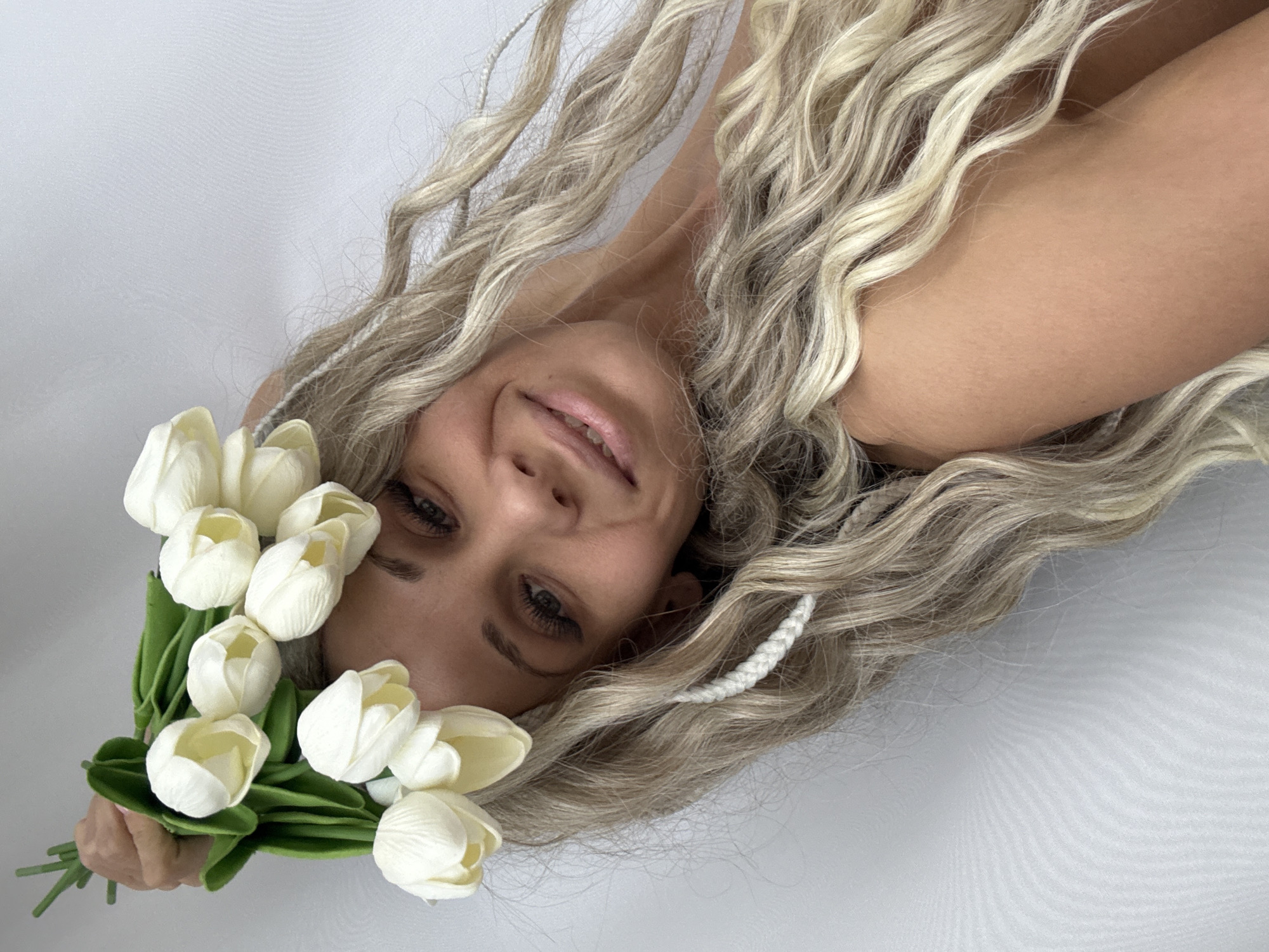 A woman with blonde curly hair is holding a bouquet of white tulips. She is lying down on a white surface. Her hair is loose and flowing around her face and shoulders. She is smiling slightly. The background is plain white. The image is well-lit and in focus.