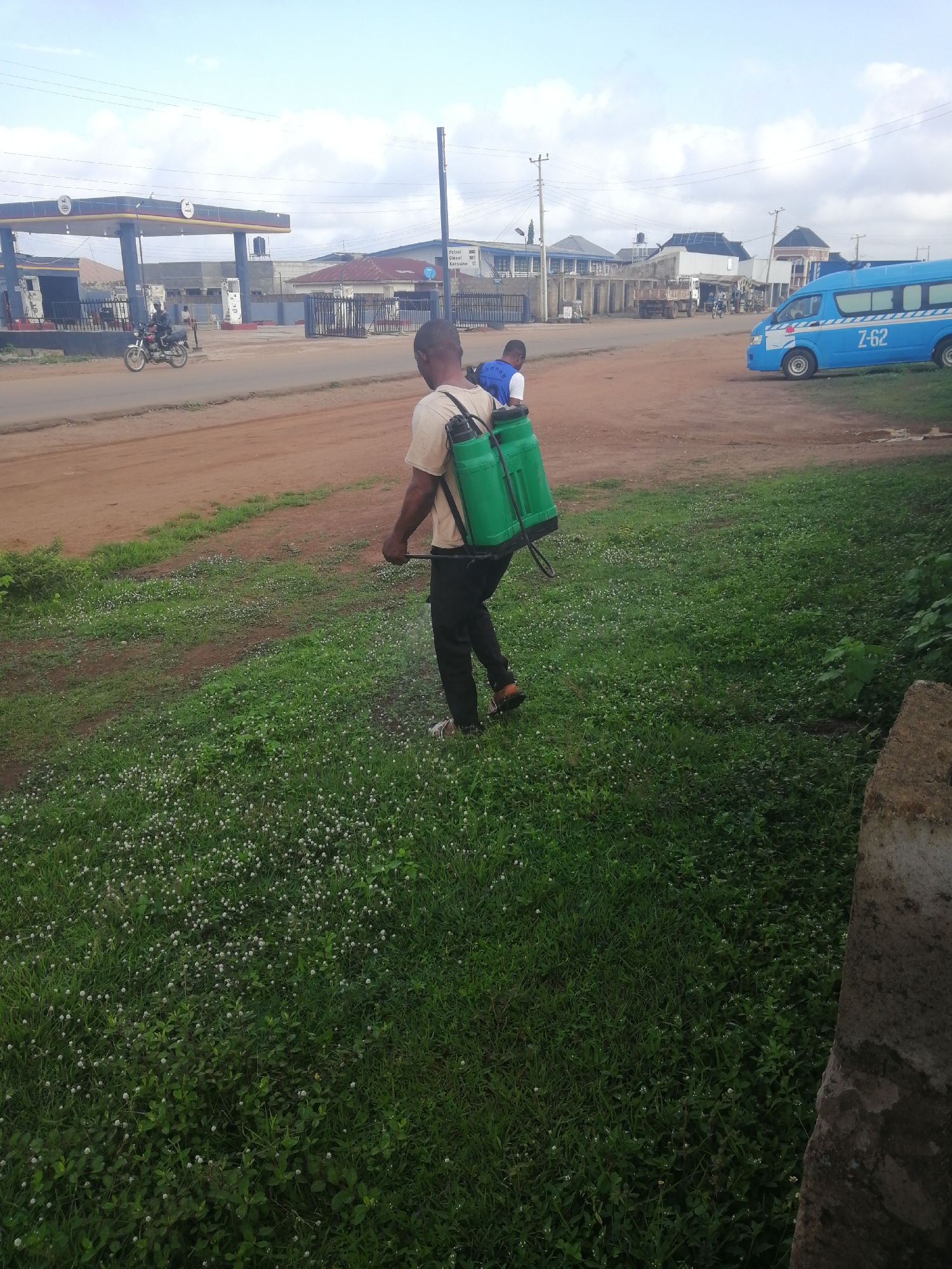 In Ajasse Ipo, Nigeria, under a cloudy sky, a man is spraying a green field with a portable sprayer. He is wearing a light-colored shirt and dark pants, with the sprayer backpack covering his back. A child is partially visible, peeking over his shoulder from within the sprayer. The field is covered in lush green grass dotted with small white flowers, with a dirt road running alongside it. In the background, a gas station with a blue and white canopy and multiple pumps is visible, along with several buildings. A blue bus with "Z-62" written on its side is parked on the road. The overall scene suggests a normal day with agricultural or maintenance work taking place near a public area.