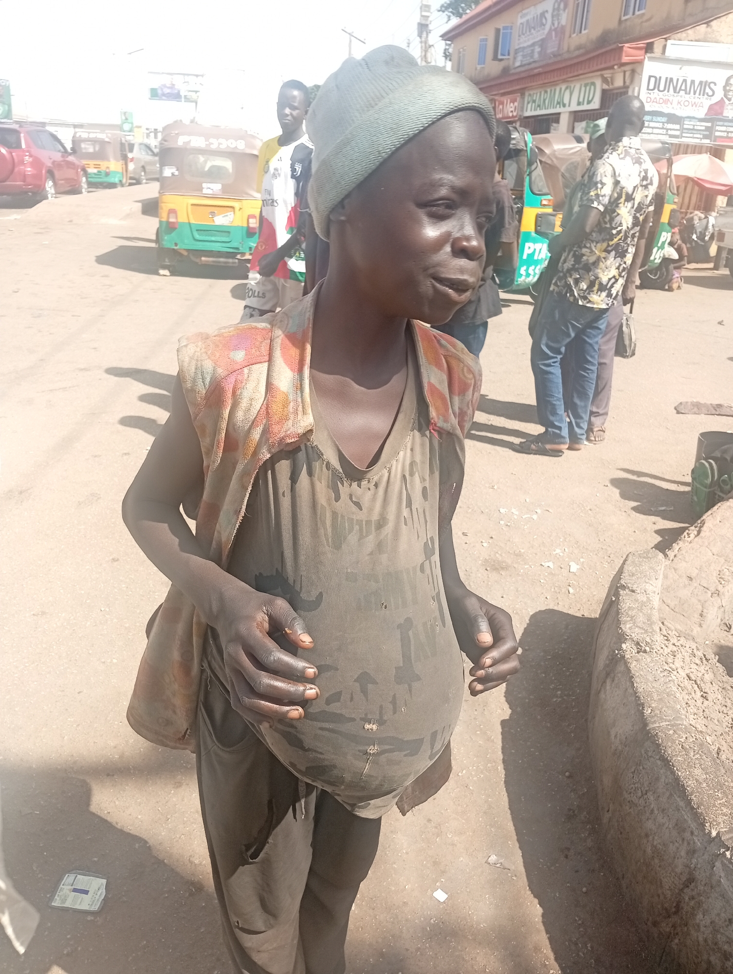 The image shows an individual with distended abdomen in an urban setting in Jos, Nigeria. The person is wearing a gray beanie, a dirty gray t-shirt, and dark pants with a torn pocket. They are standing on a paved street with several other people and vehicles visible in the background. The background includes a building with signs for "La Med Pharmacy LTD" and "DUNAMIS INT'L GOSPEL CENTRE DADIN KOWA". A three-wheeled vehicle and other cars are present in the background. The overall environment suggests a busy commercial area.
