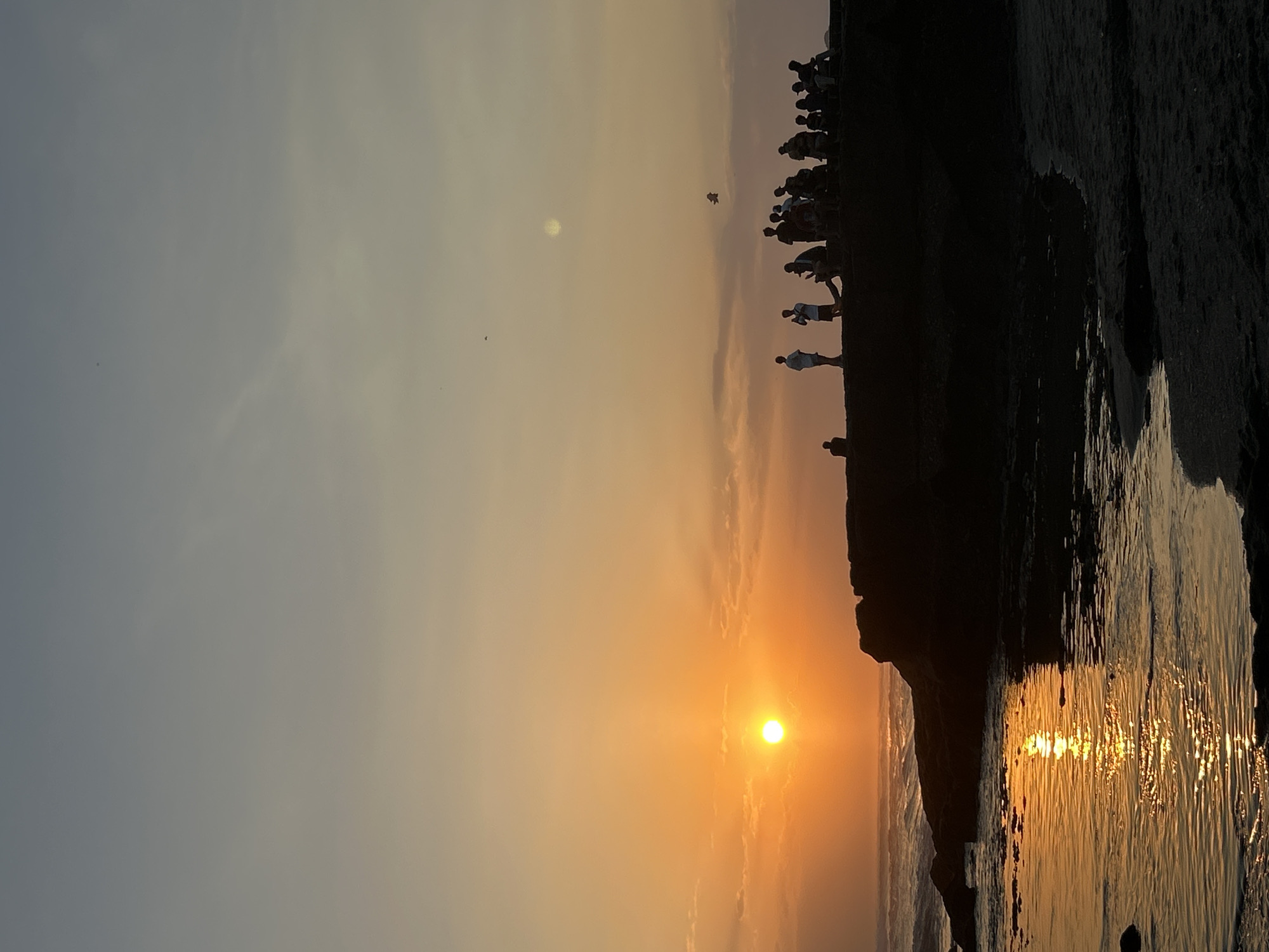 A group of people are silhouetted against the sunset on a rocky outcrop near the ocean. The sun, a bright orange orb, is low in the sky, casting a warm glow over the scene. The sky is a gradient of colors, from a pale blue at the top to a deep orange near the horizon. The ocean waves gently lap against the rocks, and a sandy beach is visible in the foreground. The people appear to be enjoying the view, with some standing and others sitting on the rocks. The location is the ocean in North Kuta, Indonesia.