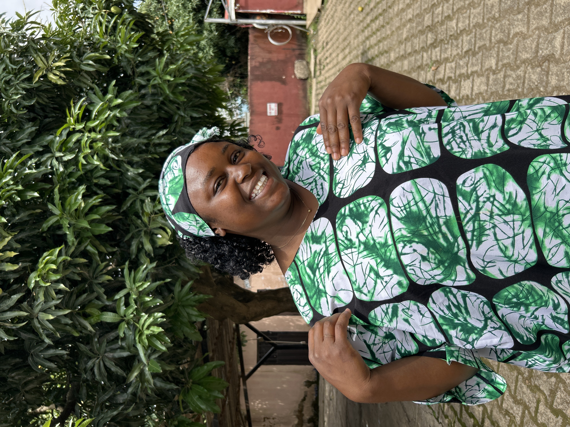 A dark-skinned female, positioned centrally and looking at the camera, stands outdoors in Jos, Nigeria. She exhibits a wide smile, revealing white teeth. Her dark, curly hair is partly covered by a green, black, and white patterned headscarf, which matches her loose-fitting top. The top features a pattern of large black ovals enclosing abstract green and white designs. A thin gold chain with a small pendant hangs around her neck, and a simple ring adorns her left ring finger. Her left hand is raised to her upper chest, while her right hand rests lower on her chest.
The background is dominated by a large tree with dense, dark green foliage, displaying visible water droplets on several leaves. To the left, a light brown building with dark wooden double doors is partially visible. To the right, a red concrete wall features a white sign with red lettering ("DANGER") and a metallic ring fixture. Further right, an open dark structure is visible. The ground is paved with grey interlocking concrete blocks, and a single yellow fruit lies on the ground to the far right.