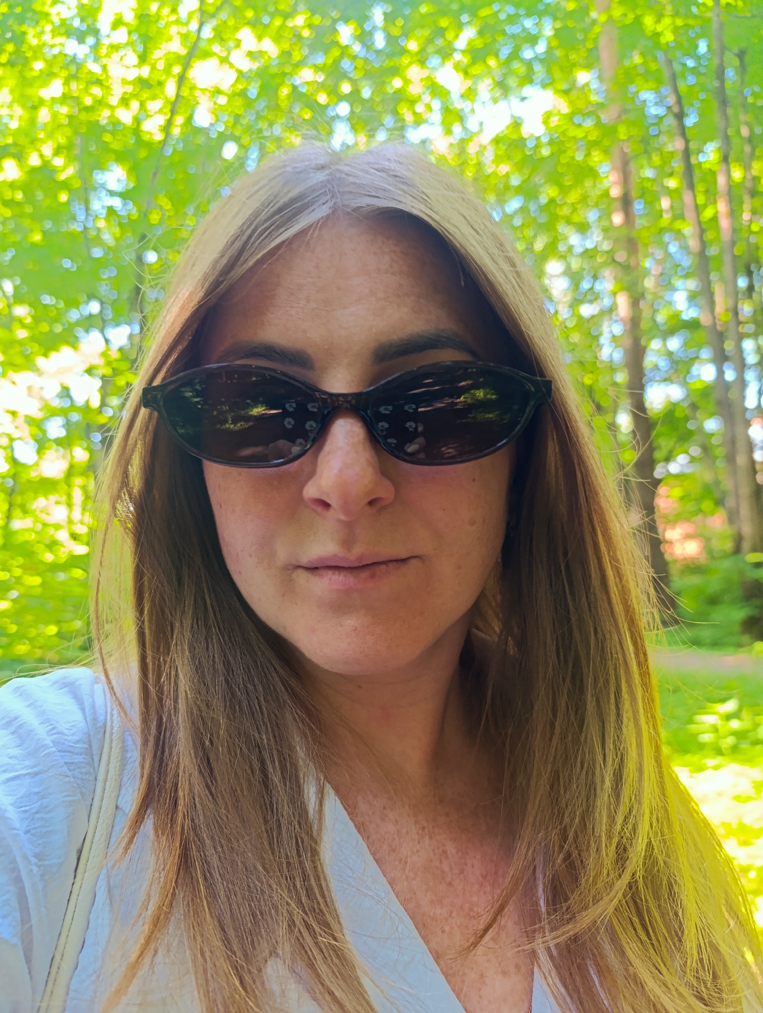 A woman is pictured from the chest up, facing the camera with a slight smile. She has fair skin with freckles and long, wavy, light brown hair. She is wearing dark sunglasses with a tortoise shell frame and a white collared shirt. The background is out of focus, showing a vibrant green forest canopy with sunlight filtering through the leaves. The image appears to be taken outdoors in a park or wooded area.