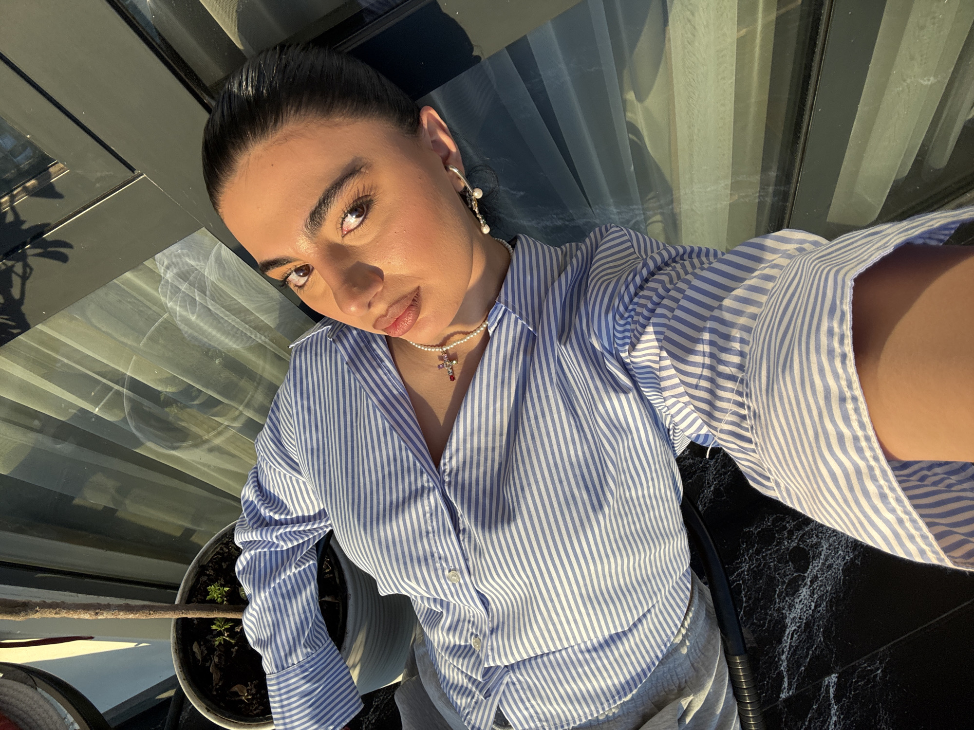 A selfie photograph shows a light-skinned woman with dark hair pulled back, wearing a blue and white striped button-down shirt and a pearl necklace with a cross pendant. She is wearing pearl earrings and is illuminated by sunlight. A potted plant is visible to her left. The background suggests an indoor space with a window or glass wall. The woman is likely in Yerevan, Armenia, though there are no concrete objects proving her location.