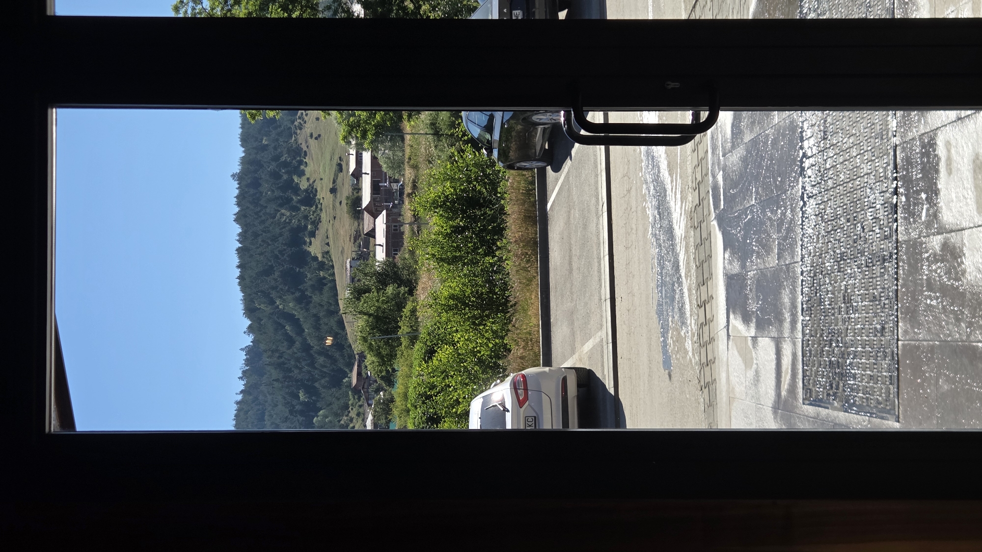The image captures a view through a doorway, looking out onto a parking lot and a landscape. In the foreground, the doorway's dark frame and a door handle are visible on the right. The ground outside appears wet, with reflections and a patterned mat at the bottom of the frame.
The parking lot contains two cars: a white sedan parked on the left and a dark sedan parked on the right. Beyond the parking lot, a row of lush green bushes and trees separates it from a village or small town. Several buildings with red roofs are nestled among the greenery. In the background, a densely forested hill rises, with a clear blue sky above. The overall scene suggests a sunny day in a rural or semi-rural setting.