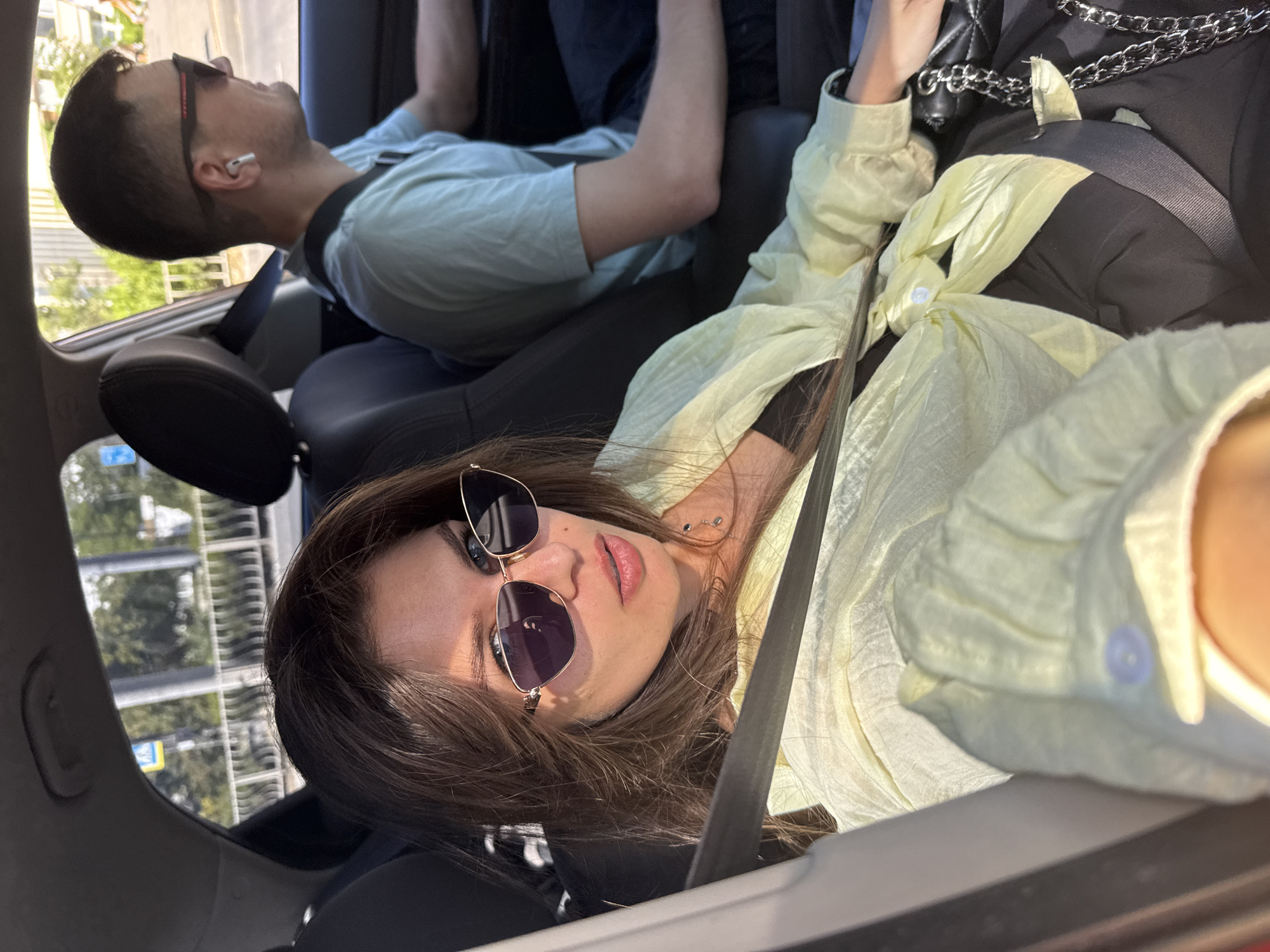 A medium shot captures two individuals inside a car on a sunny day, with trees and buildings visible through the windows. In the foreground, a woman with long brown hair and sunglasses is seated in the passenger seat, looking towards the camera. She is wearing a light yellow unbuttoned shirt over a black top and dark pants. A black purse with a silver chain strap rests on her lap. A seatbelt crosses her body diagonally. In the background, a man with short dark hair and sunglasses is driving, wearing a light blue t-shirt and earbuds. The interior of the car is dark, with the seats appearing to be black. The environment outside the car suggests an urban setting, possibly a street or highway.