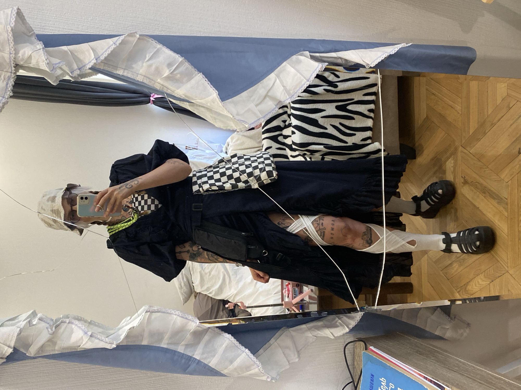 This is a vertical, eye-level, full-body mirror selfie taken by a person in a room. The person is wearing a black outfit that includes a skirt, a top with puffed sleeves, a checkered bandana, and white socks with black sandals. They are also wearing a light-colored cap. The person is holding a smartphone in their right hand, taking the picture.
The background of the image shows a bedroom with a wooden floor. A bed is partially visible, covered with blue and white bedding, and a zebra-striped blanket. A mirror reflects the person and the room. There are also some objects on a surface in the lower right corner, including books with Cyrillic text. The overall aesthetic of the image suggests a trendy, urban environment, possibly within Tbilisi, Georgia. There are no clear indicators of the time of day or weather conditions. The mood appears to be casual and self-expressive, with the person posing confidently for the camera.