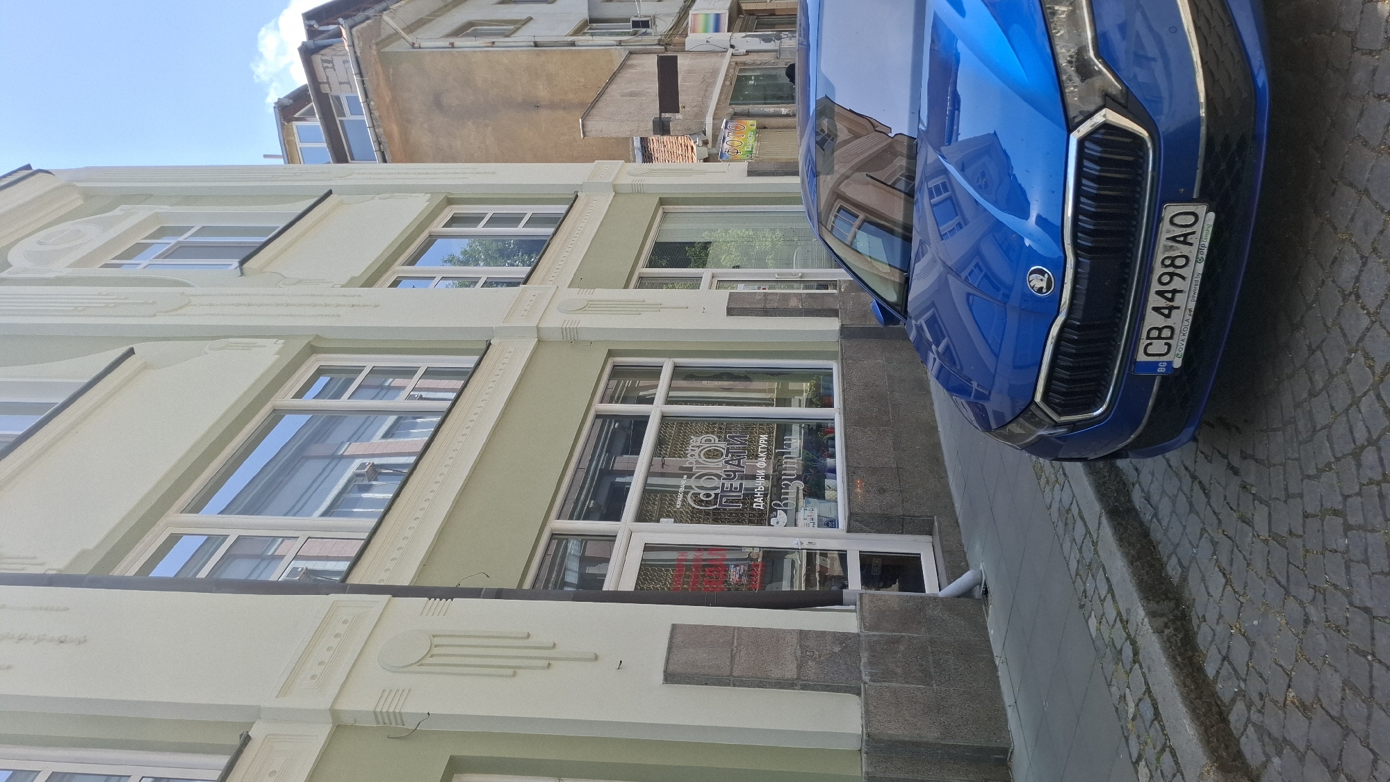 The image shows a blue car parked on a cobblestone street in front of a light green building. The building has several windows and a storefront. The storefront has text on it that reads "COLOR ПЕЧАТИ" and "DÁNSKI FAKTURI" and "Buzimku". There is also text that reads "Brother". The car has a Bulgarian license plate that reads "CB 4498 AO". The street is lined with cobblestones and there is a sidewalk next to the building. The sky is blue and there are some clouds. The overall scene is a street view in a city.
The building on the left appears to be a commercial establishment, possibly a printing shop or a business that offers related services, given the text visible on the storefront. The architectural style of the building suggests it's in an older part of the city. The presence of a blue car, a common mode of transportation, and the cobblestone street are characteristic of many European cities. The license plate clearly indicates the car is registered in Bulgaria. The context of Plovdiv, Bulgaria, adds a specific geographical element, suggesting the photo was taken in this historic city.