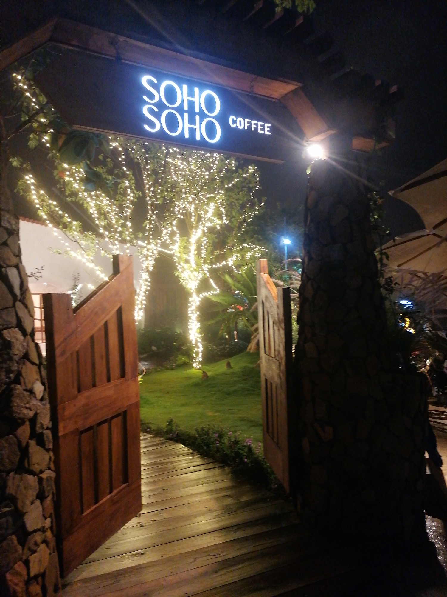 The image displays the inviting, illuminated entrance of "SOHO SOHO COFFEE" at night, likely situated in Da Lat, Vietnam. A large, two-leaf wooden gate stands open, revealing a garden beyond. The gate is rustic in design, with vertical wooden panels, and is framed by substantial pillars constructed from rough-hewn stones. A curved wooden pathway, made of horizontal planks, leads from the gate into the verdant garden.
The garden is dominated by several trees extensively decorated with numerous small, warm-toned string lights, creating a sparkling and ethereal ambiance against the dark night sky. Patches of green grass are visible beneath the trees and alongside the wooden path, complemented by various plants and shrubs. A white building with a visible window can be seen in the background to the left, while part of a large outdoor umbrella or canopy is discernable on the far right, suggesting an outdoor seating area.
Above the entrance, a prominent, rectangular sign glows brightly with white, sans-serif text. It reads "SOHO SOHO" stacked vertically, with "COFFEE" in smaller letters to the right of the lower "SOHO". A strong light source behind the right stone pillar creates a visible lens flare. The overall atmosphere is warm, magical, and welcoming, characteristic of an evening coffee shop. No people are present in the scene.