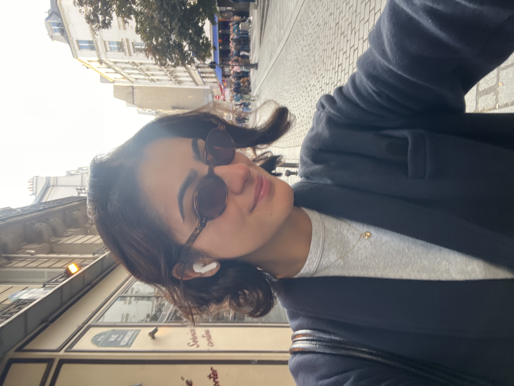A medium, tilted shot depicts a woman wearing sunglasses, an earbud, a gray shirt, and a dark coat, on a street in Paris, France. In the background, there are buildings and people. A sign with "Rue de Verberie" is visible. The ground is brick.