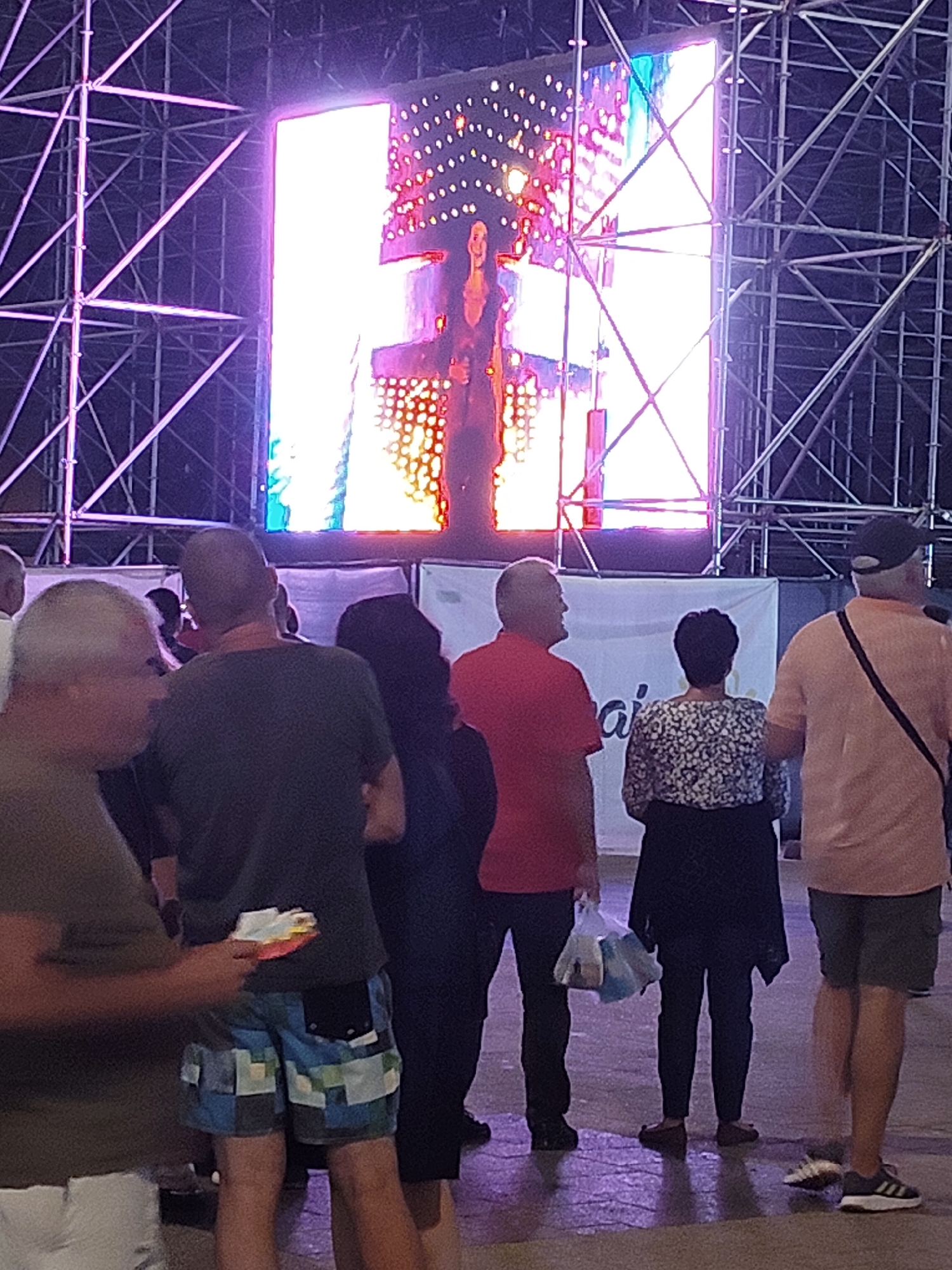 The image depicts an outdoor event with a large LED screen displaying a performer. In the foreground, several individuals are visible, seemingly attending the event. To the left, a man wearing a khaki t-shirt and white shorts is partially visible. Next to him, a man in a dark t-shirt and patterned shorts holds a small stack of colorful papers. Further to the right, a person in a red shirt stands facing away from the camera, carrying a plastic bag. Behind them, a woman with short dark hair wears a patterned top and dark skirt. To the far right, a man in a peach-colored t-shirt and dark shorts stands with a bag strap over his shoulder. The background features a metal scaffolding structure supporting the LED screen, which shows a bright, pixelated image of a woman. The ground appears to be paved and wet, reflecting the purple and white lights from the screen and scaffolding. The scene suggests a concert or performance taking place in a public area, possibly a festival or outdoor venue. Based on available information about similar events and common outdoor venues in Constanța, Romania, this could be an event held at the Constanța Exhibition Center or a similar open-air space.