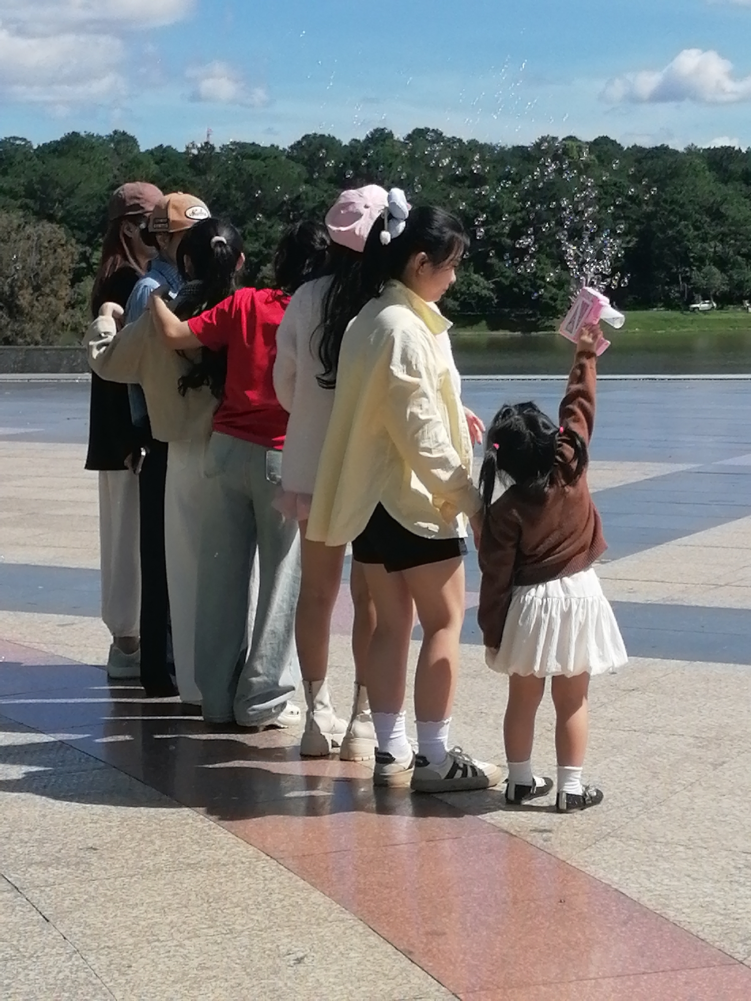 A group of people, including children and adults, are standing in a line outdoors on a paved surface. The children are holding a pink bubble machine and blowing bubbles. In the background, there are trees and a body of water. The people appear to be dressed casually, with some wearing hats and others wearing light-colored clothing. The weather appears to be sunny with some clouds in the sky. The setting suggests a public park or recreational area in Da Lat, Vietnam, likely near Xuan Huong Lake or a similar waterfront location due to the presence of water and trees characteristic of the city's landscapes. The ground is paved with textured tiles, and shadows are cast from the people and the light source, indicating it is daytime.