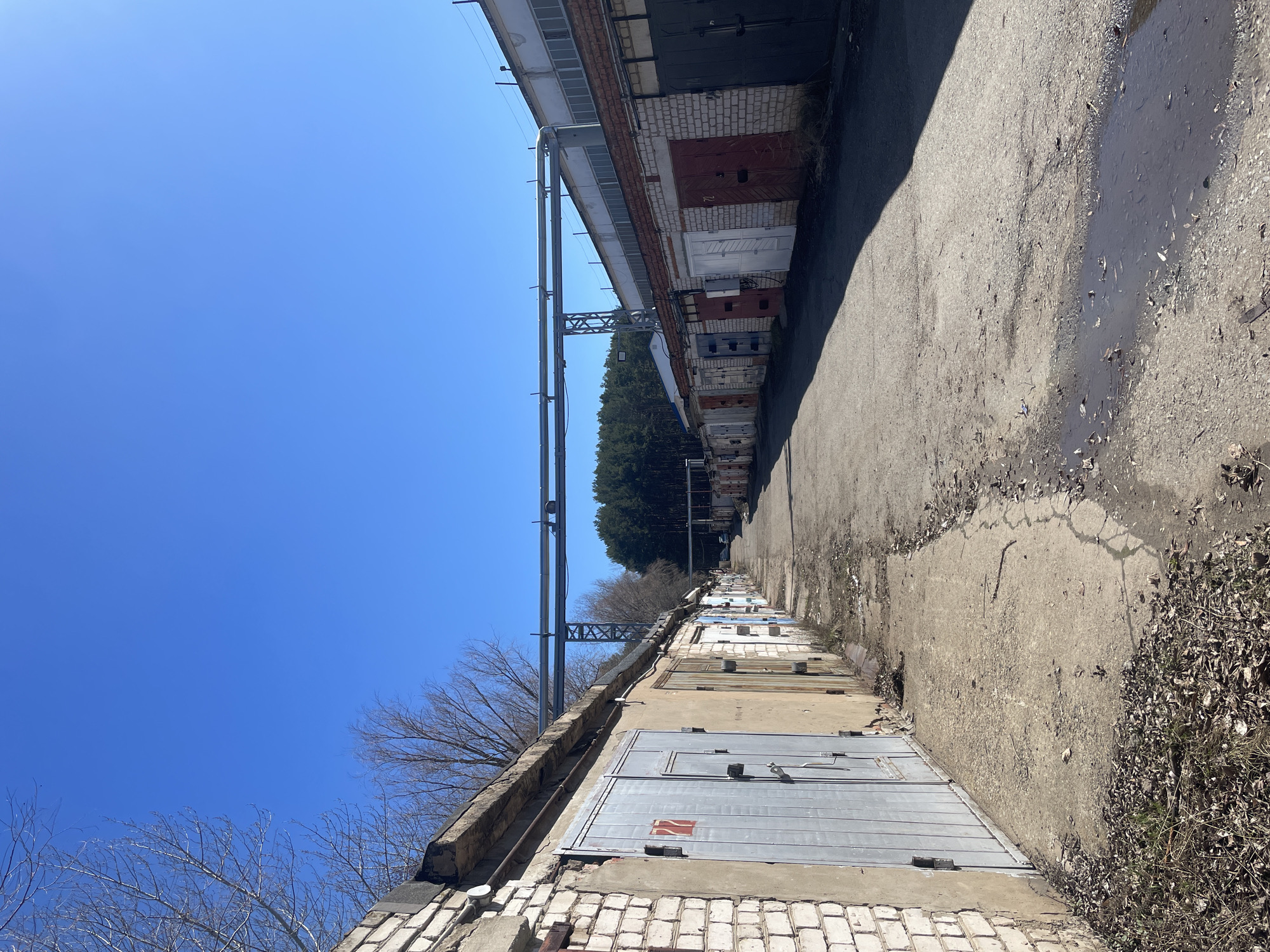 The image shows a row of garage doors along a cracked concrete alleyway in Chaikovsky, Russia. The garages are constructed of brick, with metal doors of varying colors and conditions. Some have visible numbering. A metal pipe runs along the top of the garages, supported by metal beams. Beyond the garages on the opposite side of the alley is a line of taller brick structures, possibly more garages or industrial buildings. The alleyway is cracked and uneven, with patches of water and debris. A line of trees is visible in the distance beyond the taller structures. The sky is a clear blue. The light suggests it is daytime. There are no people visible in the image.