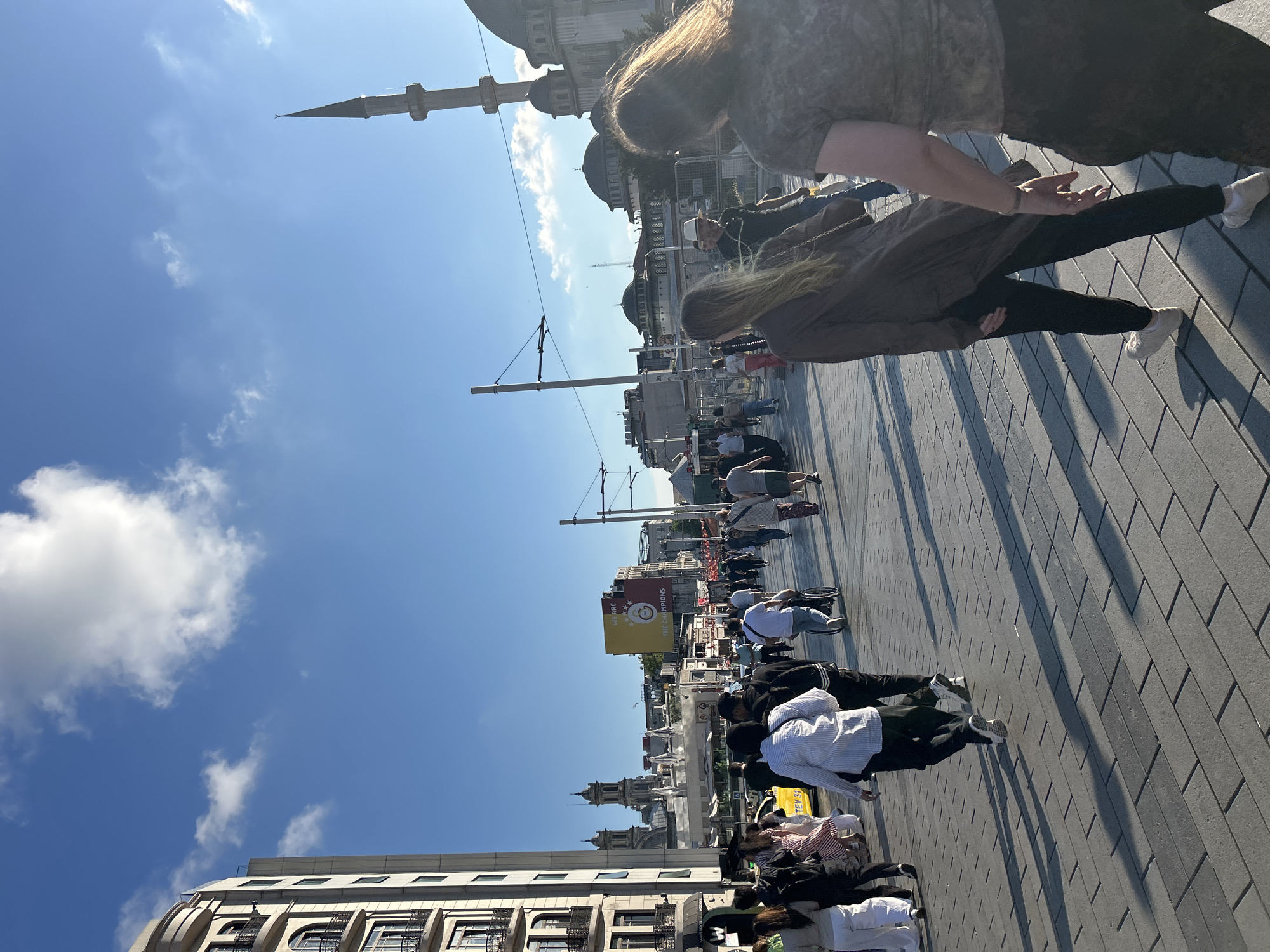 The image depicts a street scene in Beyoğlu, Turkey, under a bright, partly cloudy sky. A diverse group of people are walking on a wide, paved pedestrian walkway. Many are walking towards the viewer, while others are moving away. Some individuals are in the foreground, with their backs or sides to the camera.
Notable elements in the background include modern buildings on the left, with balconies and multiple windows. Further down the street, a large yellow billboard displays the Galatasaray S.K. logo and the text "WE ARE THE CHAMPIONS." A minaret of a mosque is visible to the right, along with dome structures of a mosque complex. A yellow taxi is parked on the street to the left. Overhead wires and street lights are present, suggesting an urban environment with public transportation or street infrastructure. The ground is paved with large, rectangular grey tiles laid in a herringbone pattern, casting long shadows from the people and structures. A person in a wheelchair is also visible among the pedestrians. The overall atmosphere suggests a busy, public urban space during daylight.