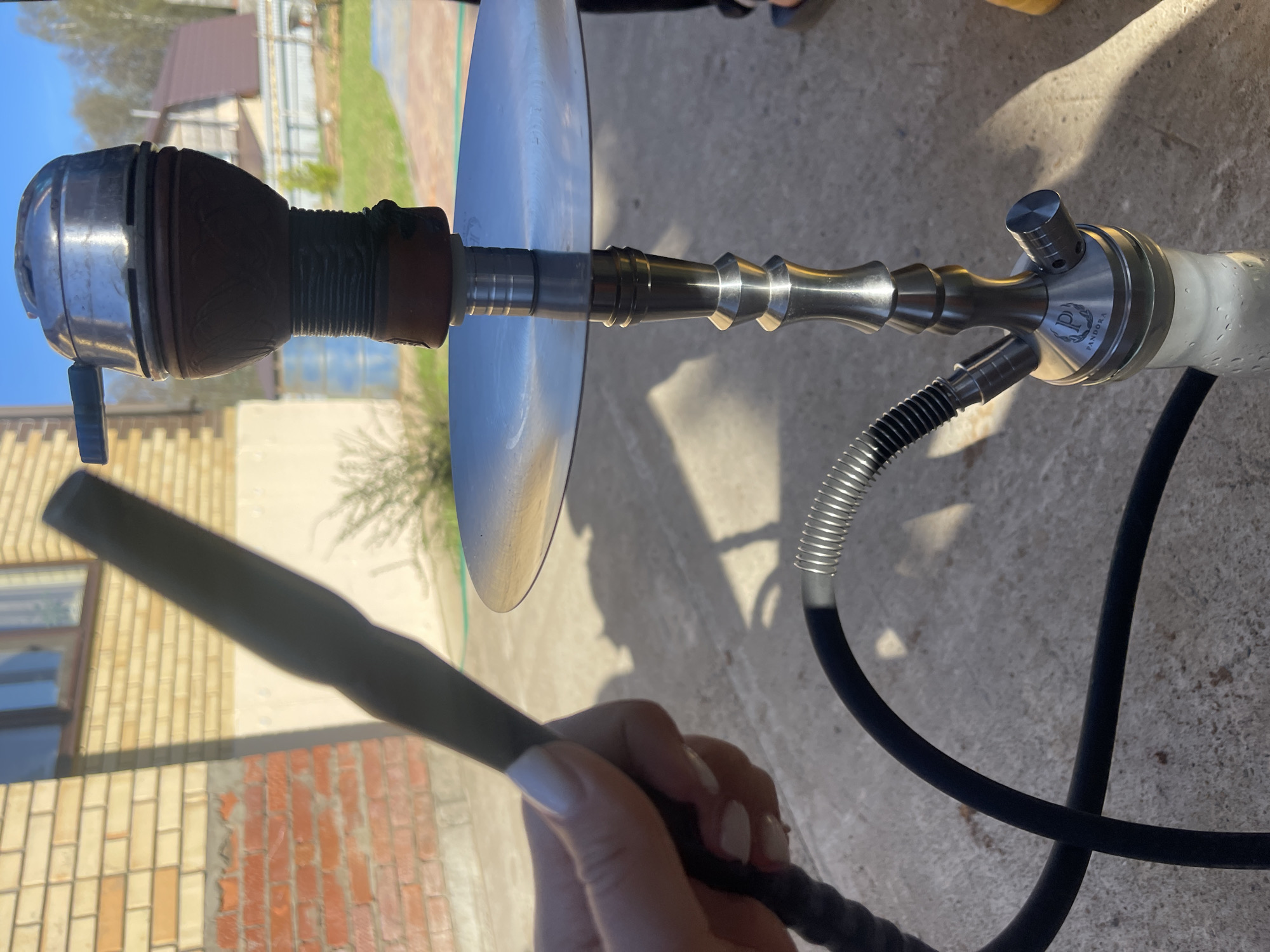 The image shows a hookah set up outdoors in Chaikovsky, Russia. The hookah consists of a charcoal holder, a metallic wind cover, a metallic ash tray, a decorated central shaft, and a glass base with a "Pandora" logo. A hand holds the black mouthpiece of the hose. The background includes a building with light brick and windows, vegetation, and a concrete surface. The scene is well-lit, suggesting it is daytime.
