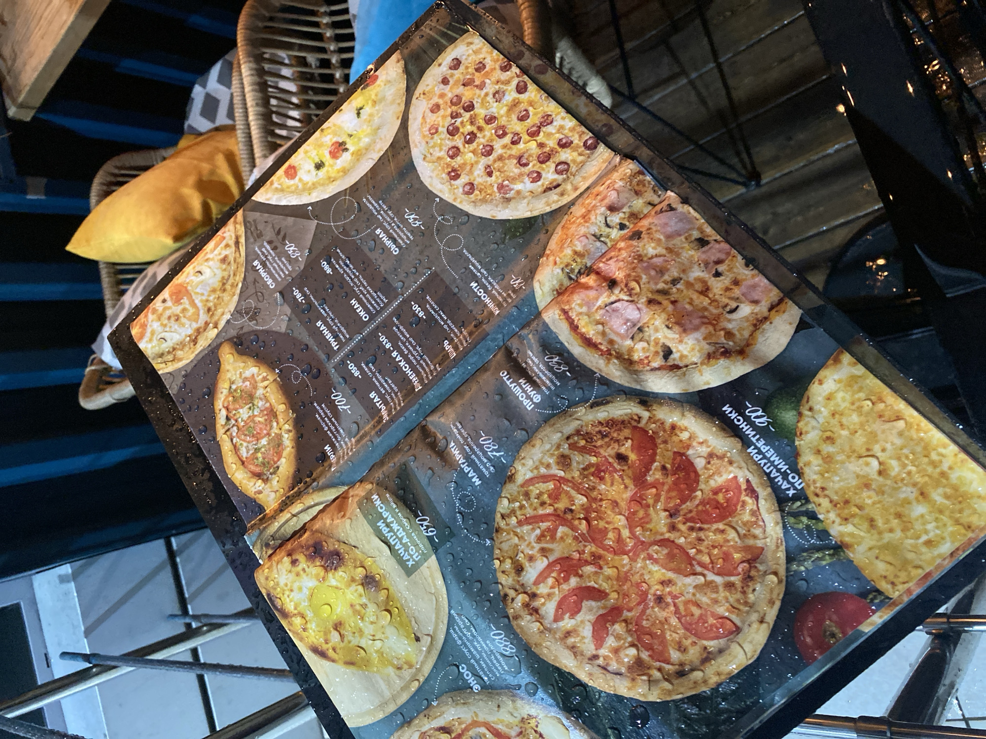 The image shows a menu on a table at a cafe or restaurant, possibly in Sochi, Russia, based on the Cyrillic script used. The menu primarily features different types of khachapuri and pizza, with descriptions of ingredients and prices in Russian. The table surface has water droplets on it, suggesting recent rain or cleaning. The background shows hints of outdoor seating with blue accents and a woven chair. The focus is on the menu and the food items listed.