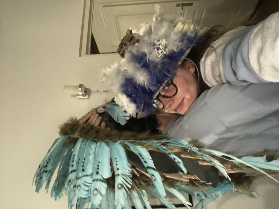 A person wearing glasses is positioned slightly tilted to the side. They are wearing a blue and white feathered hat with a toy animal on top, along with a brown and blue feathered garment or accessory. The background consists of a white wall, a light fixture, and the edge of a door or mirror. The person is wearing a light blue sweatshirt.
