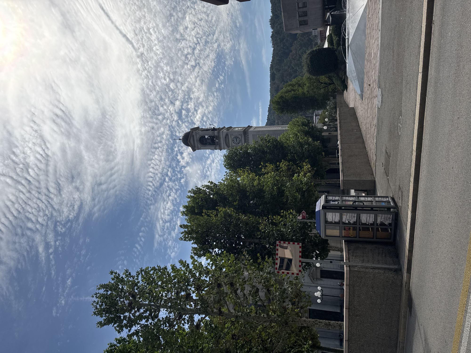 The image depicts a street scene in Chiasso, Switzerland, with a prominent clock tower in the background. The clock tower, made of stone, has a bell at the top and a clock face with Roman numerals. It is surrounded by lush green trees. In the foreground, a glass-enclosed structure, possibly a phone booth or a small kiosk, is visible. The kiosk has shelves inside filled with what appear to be books. A reflective mirror, framed with red and white checkered tape, is mounted on a pole to the left of the kiosk. The street itself is paved with asphalt, marked with a yellow line. Beyond the kiosk and trees, a modern building with geometric shapes and a fountain can be seen. The sky is blue with scattered, wavy clouds. The overall impression is an urban environment with a mix of historical and modern elements.