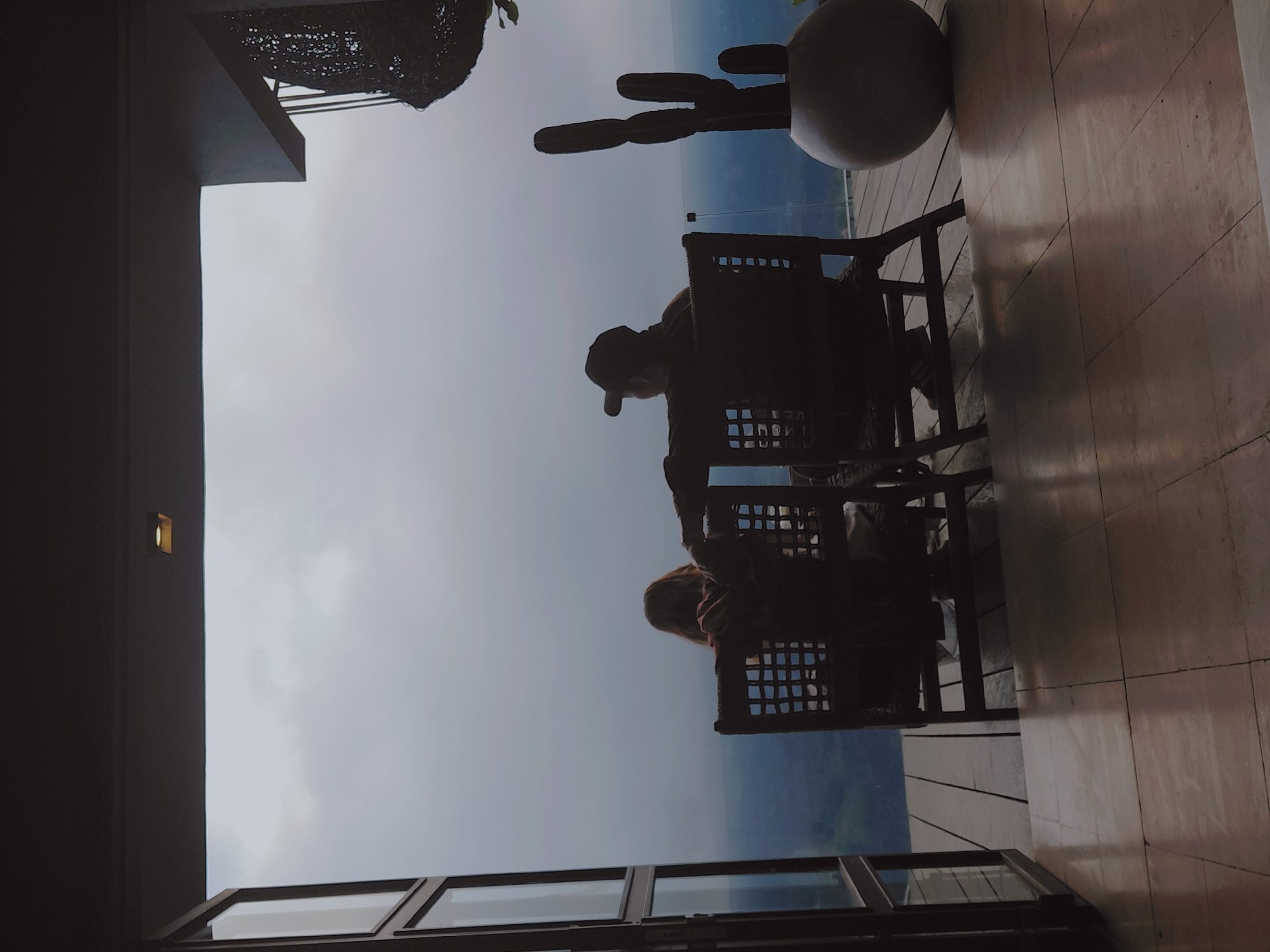 In Kintamani, Indonesia, two individuals are seated in chairs on a wooden deck, facing away from the camera and towards a hazy, expansive view. The person on the left, with long, light-colored hair, is wearing a reddish garment. The person on the right, wearing a baseball cap, has their arm resting on the back of the other person's chair. A large, round planter containing a cactus is positioned to the right of the chairs, and a decorative woven object hangs from the ceiling above. The scene is framed by an open doorway on the left and a tiled floor in the foreground. The sky is overcast, creating a muted, atmospheric backdrop.