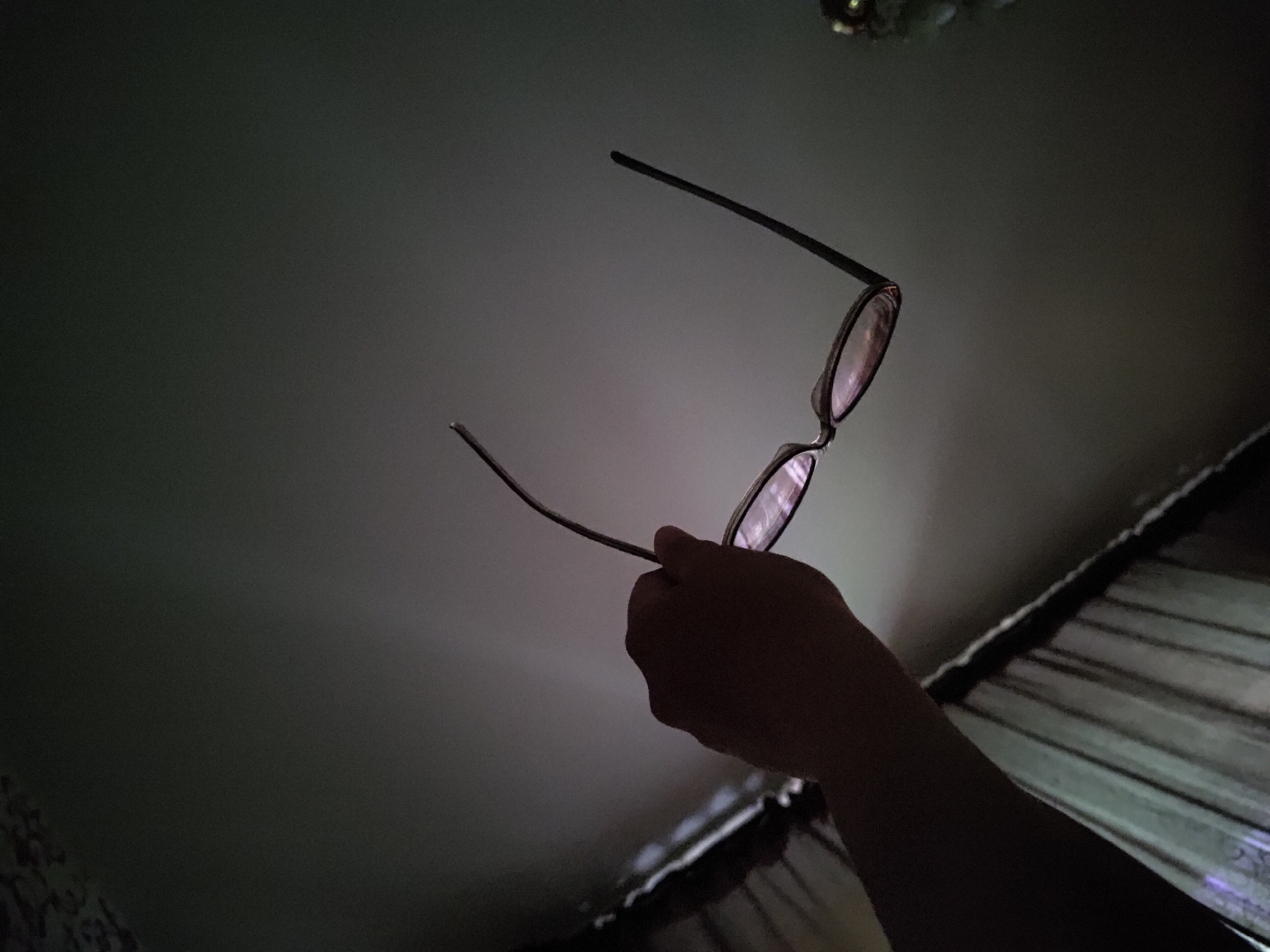 A human hand holds a pair of glasses against a dark background. The location is Марковский, Russia. The glasses are held up with the lenses reflecting light from the ceiling or a nearby light source. The arm holding the glasses extends downward to the lower right of the frame. The background is poorly lit, with portions possibly showing a ceiling.