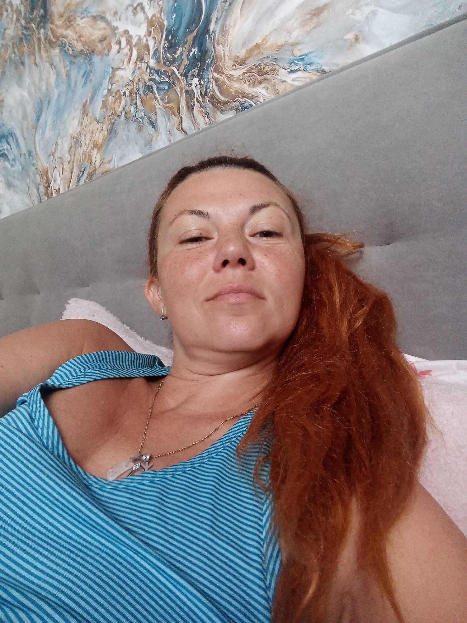 A medium shot photograph shows a woman with reddish-brown hair lying on a bed. She is wearing a blue and white striped top and a necklace with a pendant. The background consists of a gray headboard and a wall with an abstract painting. The woman is looking directly at the camera.