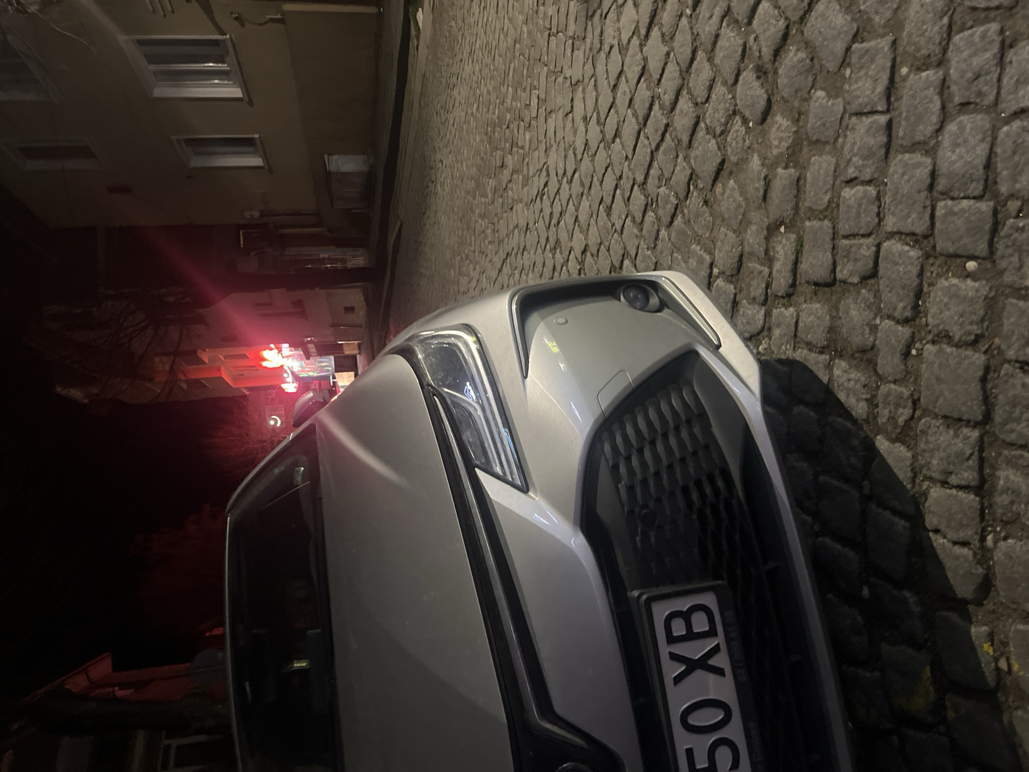 The image shows the front of a silver or white car parked on a cobblestone street. The license plate is partially visible. In the background, there is a building with windows and lights. The overall lighting suggests it is nighttime. The partial license plate reads "50 XB". The presence of cobblestones, along with other indicators (building style, partially visible license plate) strongly suggests the location may be in an urban area of Goce Delcev, Bulgaria.