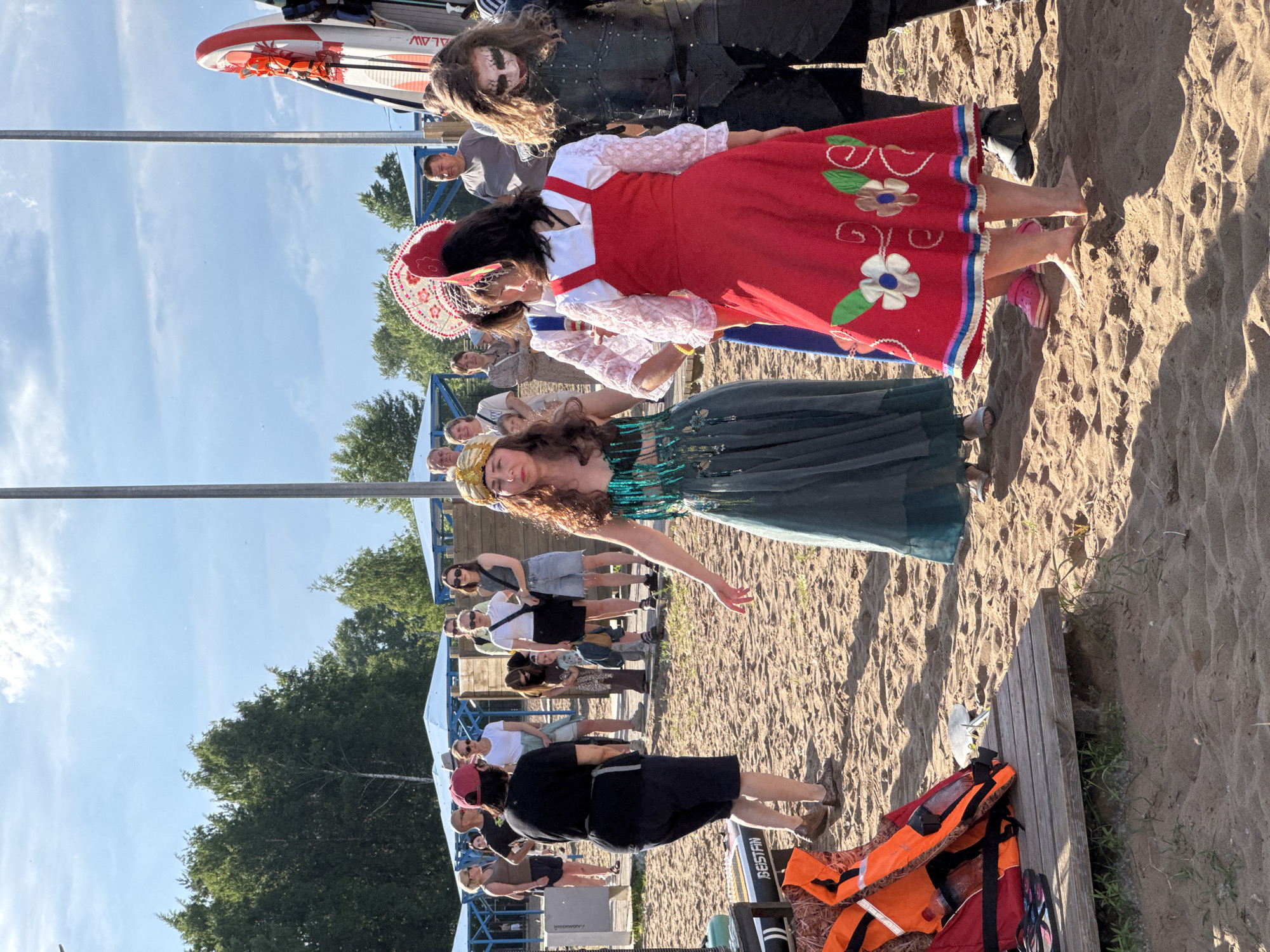 The image depicts an outdoor gathering on a sandy beach, likely near the city of Chaikovsky, Russia, given the context provided. In the foreground, a woman in a teal, sequined dress and a golden headdress is seen dancing. To her right, another woman wears a traditional red dress adorned with floral embroidery and a white, elaborate headpiece. A third person, to the far right, is dressed in a dark, armor-like costume with white face paint. Several other individuals, dressed in casual attire, are observing the scene in the background. Life jackets and a wooden platform are visible on the sand. The environment includes trees and a bright, partly cloudy sky.