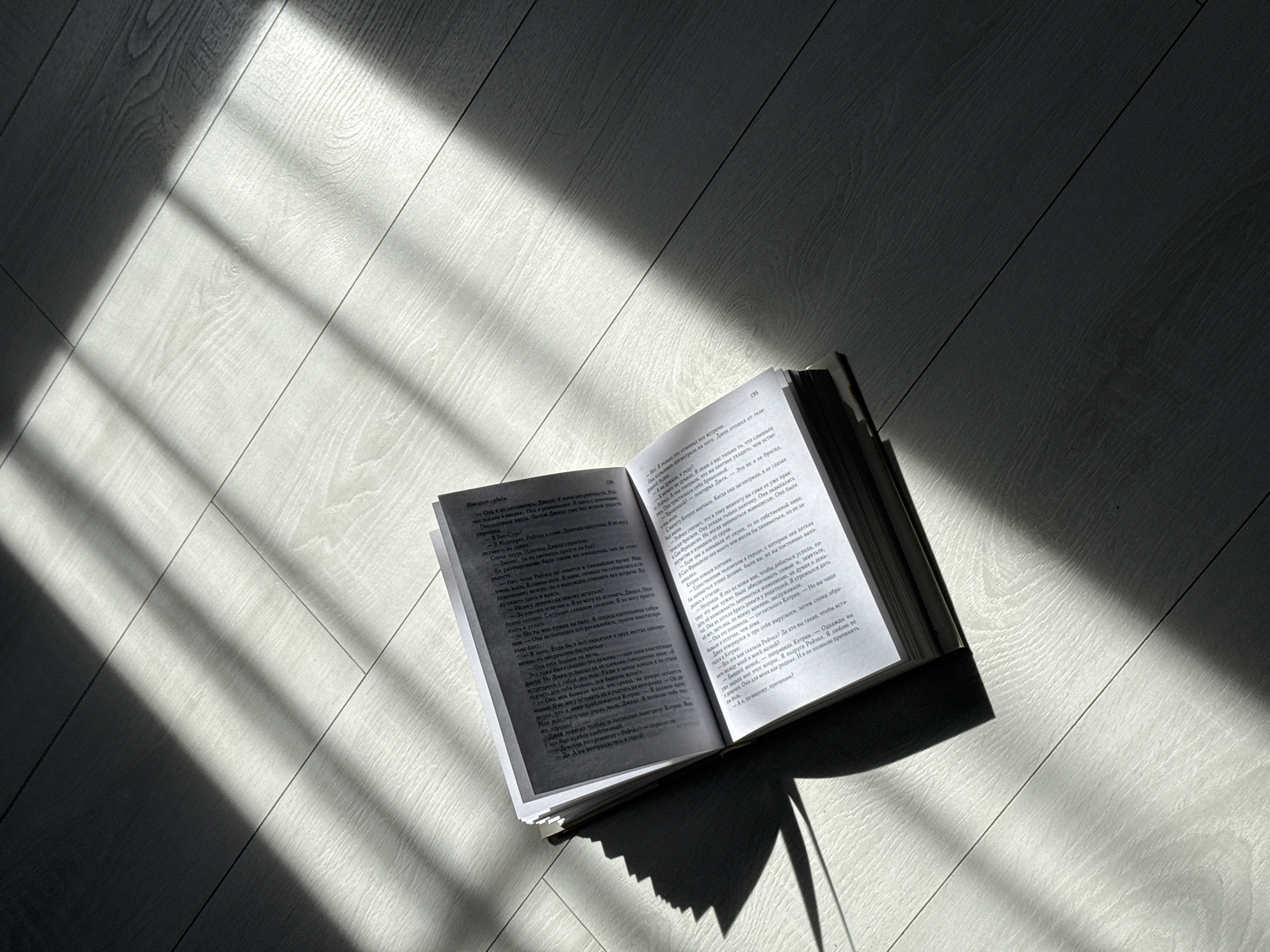 An open book rests on a light-colored wooden floor, illuminated by strong, angled sunlight. The light casts distinct, parallel shadow stripes across the floor and the book, suggesting direct sunlight filtering through a window, likely in the morning or late afternoon. The wooden floor has a visible grain and plank structure.
The book is open to pages 194 and 195. The text is printed in Cyrillic script, identified as Russian. The title "Поворот судьбы" (meaning "Turn of Fate" or "Twist of Fate") is visible at the top of page 194. The visible content on both pages appears to be narrative prose interspersed with dialogue, indicated by leading dashes. No people are present, and the scene evokes a quiet, contemplative atmosphere.