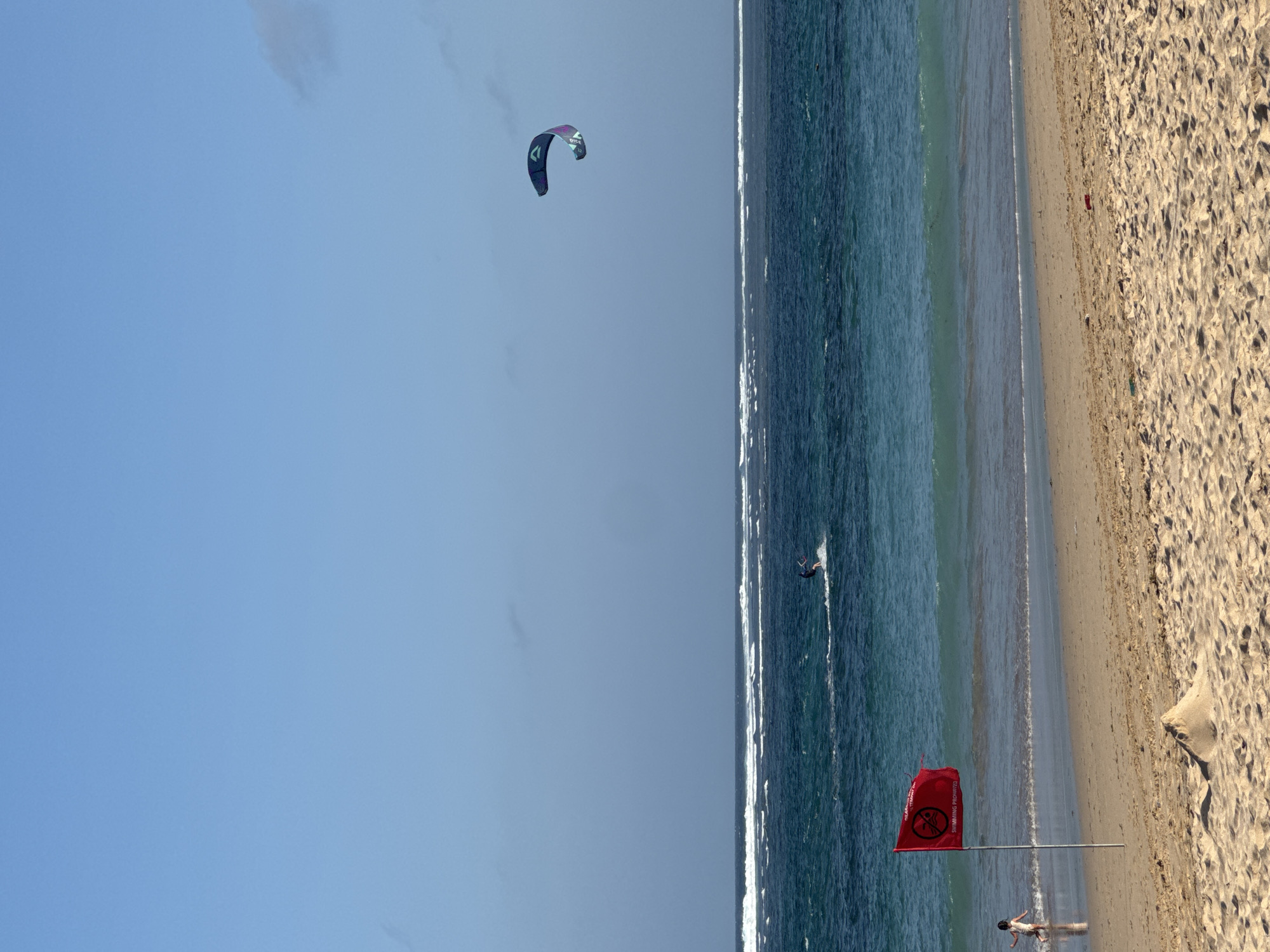 This image depicts a beach scene in Kuta Selatan, Indonesia, under a clear blue sky. The foreground is dominated by a sandy beach with a small sandcastle. A red flag with a white circle and diagonal line indicating "swimming prohibited" flies on a pole to the left. In the shallow water near the shore, a child in a white swimsuit is visible. Further out in the turquoise ocean, waves break, and a kitesurfer is engaged in the sport. In the distance, another kitesurfer is also visible. A large, colorful kite hovers in the sky above the ocean. The water is a vibrant blue-green, with white crests on the waves. The overall scene conveys a day at the beach with recreational activities, but with a clear warning against swimming.
