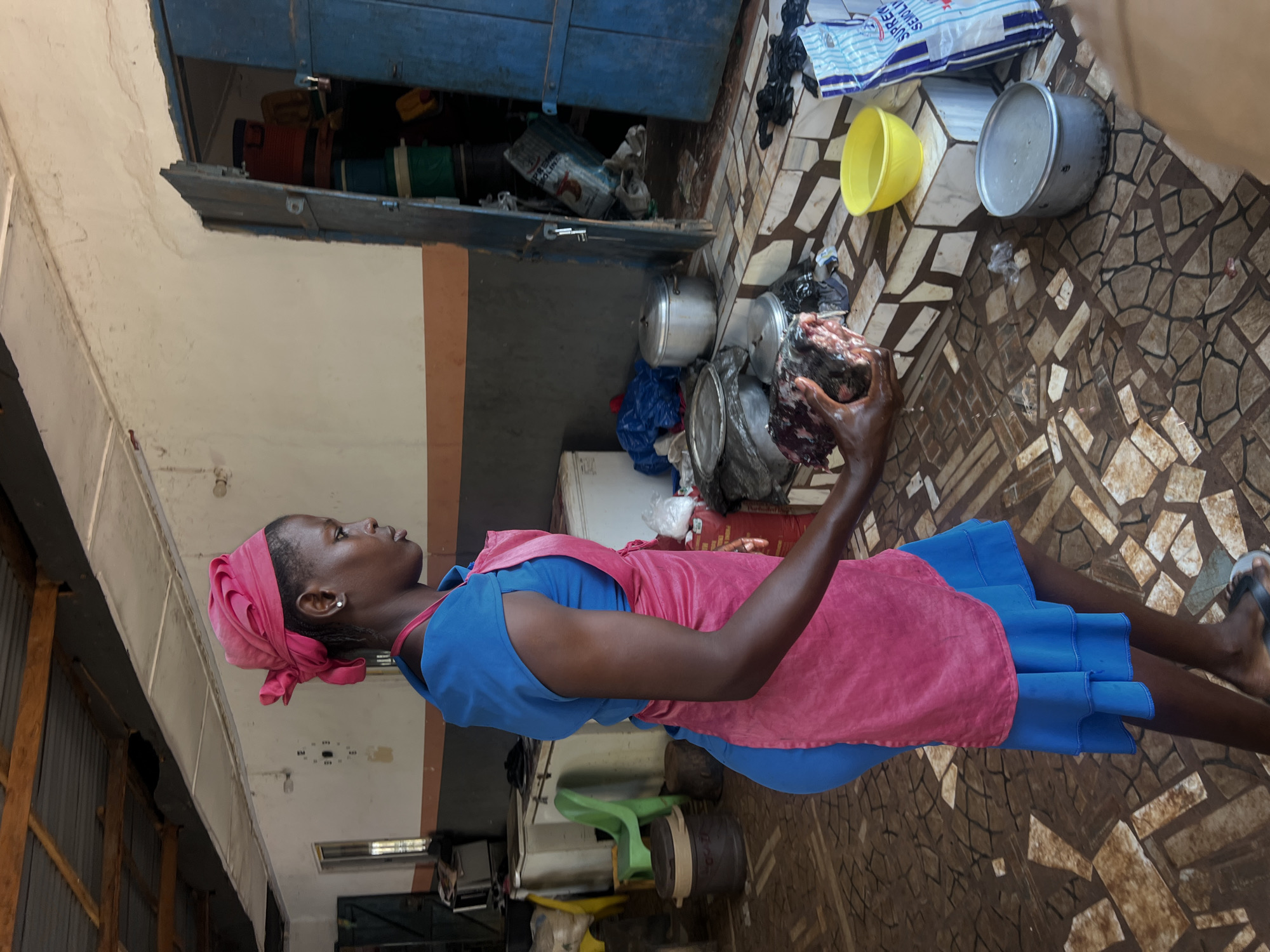 A side view shows a young Black woman wearing a blue dress and a pink apron. She has a pink headscarf tied around her head. She is holding a piece of raw meat in her right hand. She is standing on a tiled floor in what appears to be an outdoor market or food stall. The background shows a covered area with wooden beams, a wall with an orange stripe, and an open doorway revealing stacked items. There are various containers, pots, and bags scattered around the scene. Water is visible on the floor. The overall impression is that of a busy, working environment in Nkpor, Nigeria.
