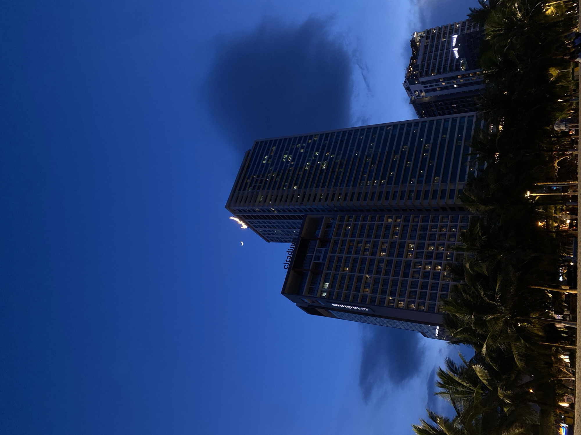 The image depicts a twilight scene in Nha Trang Ward, Vietnam, featuring two modern high-rise buildings silhouetted against a deep blue sky. The sky is partially obscured by dark, wispy clouds, one of which forms a large, amorphous shadow in the upper left quadrant of the frame. A sliver of a crescent moon is visible in the sky between the two buildings.
The buildings themselves are tall and rectangular, with many windows illuminated by interior lights, suggesting occupancy. One building, positioned closer to the viewer and angled more prominently, displays the illuminated text "Citadines" vertically along its side. The other building, further in the background, also shows illuminated windows.
In the foreground, the dark silhouettes of palm trees create a tropical ambiance, with their fronds overlapping and obscuring parts of the buildings and the background. The lighting suggests it is either dusk or dawn, with the artificial lights of the buildings and distant illumination creating a contrast with the natural twilight. The overall mood is serene and urban, with a touch of natural beauty provided by the palm trees and the sky. There are no people clearly discernible in the image, but the illuminated windows imply human presence within the buildings.